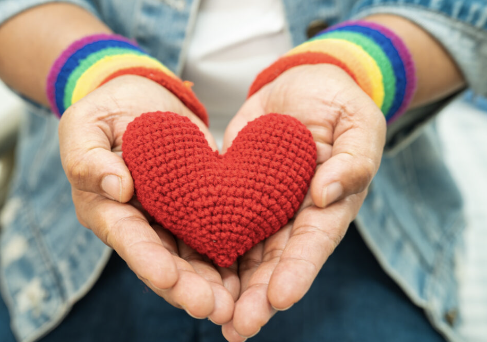 Cupped hands wearing rainbow-striped wristbands holding a red crocheted heart
