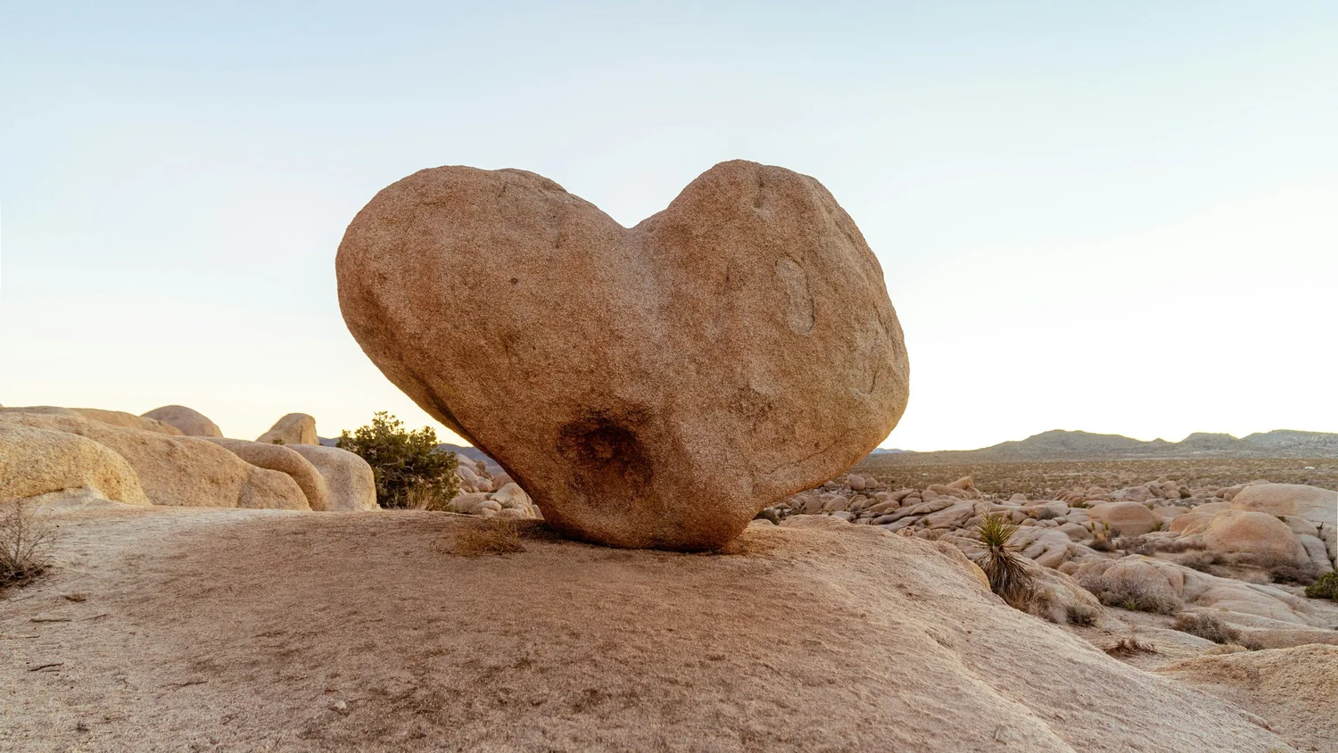 Large heart-shaped boulder resting on smooth rock in a desert landscape under a pale sky