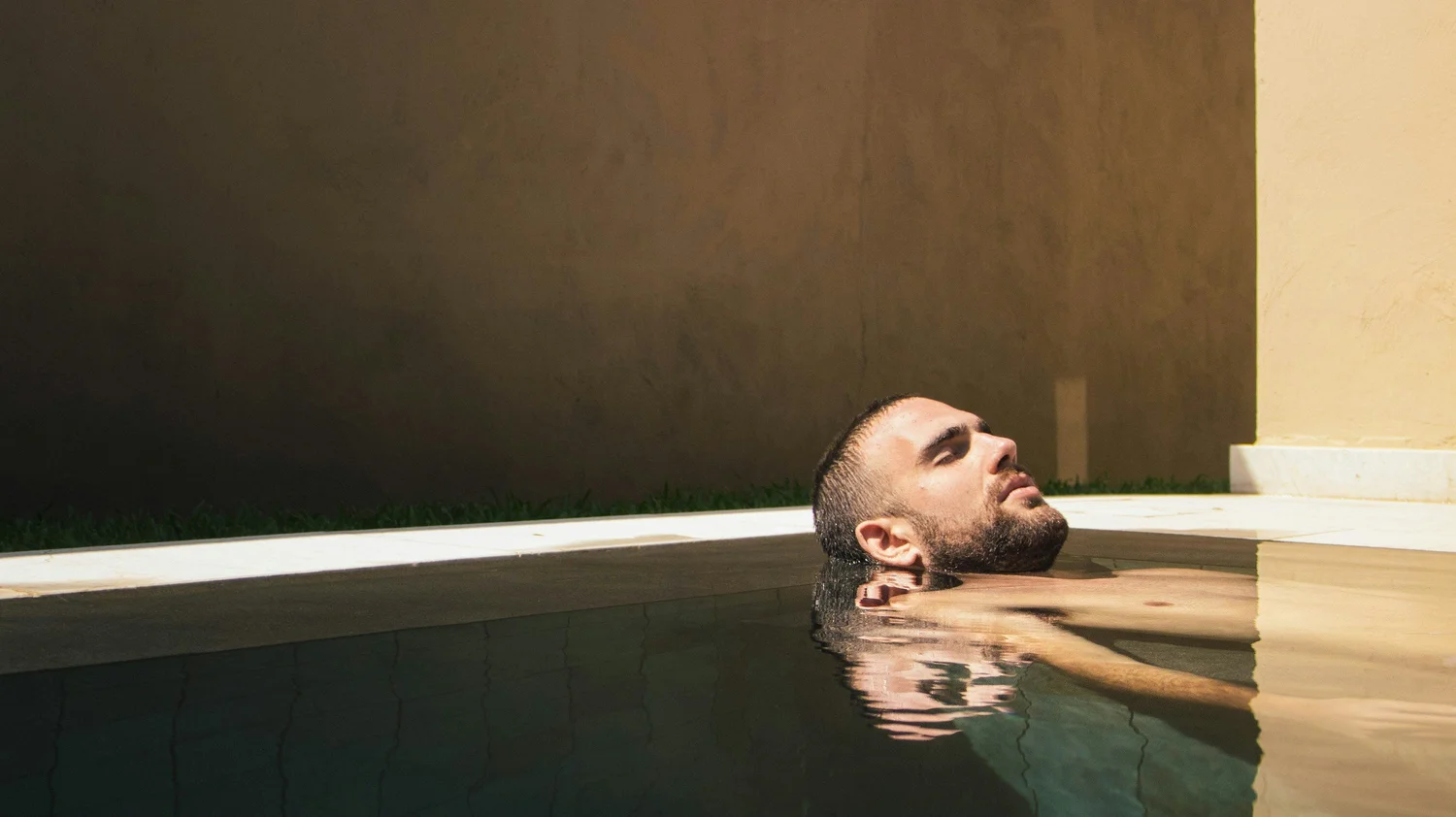 A person reclining in a pool with head tilted back and eyes closed in bright sunlight