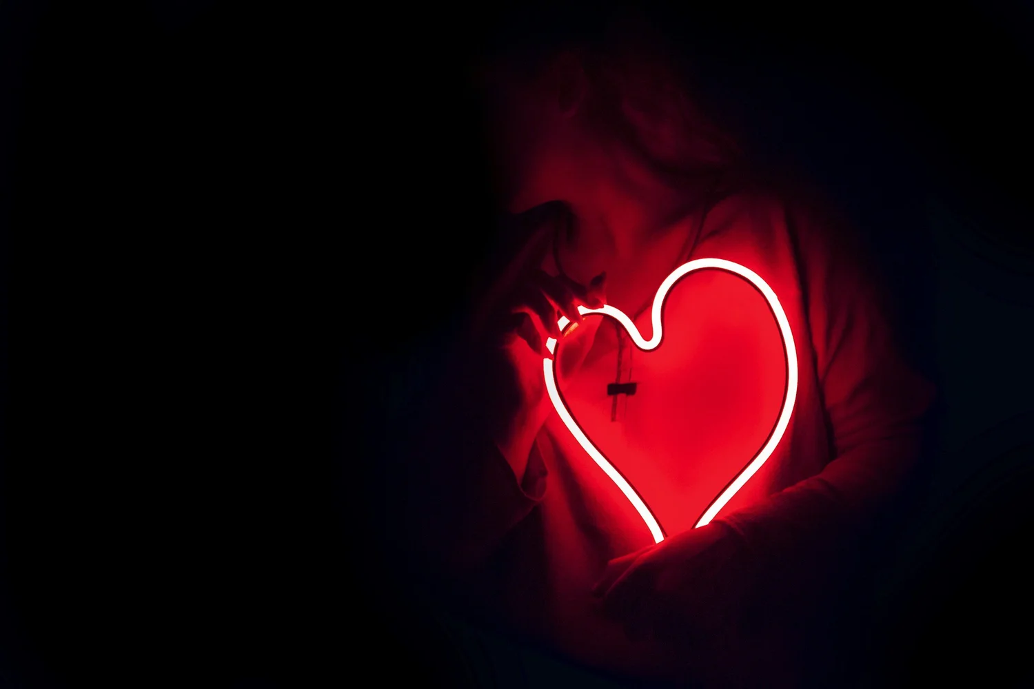 A person in darkness holding a glowing red neon heart close to their chest