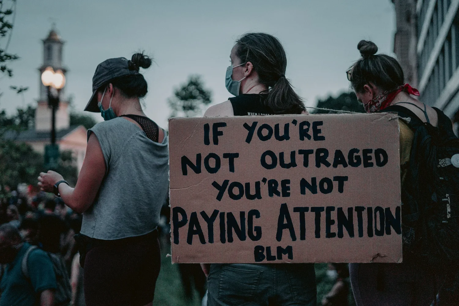 A masked person in a crowd holds a cardboard sign: "If you're not outraged you're not paying attention"