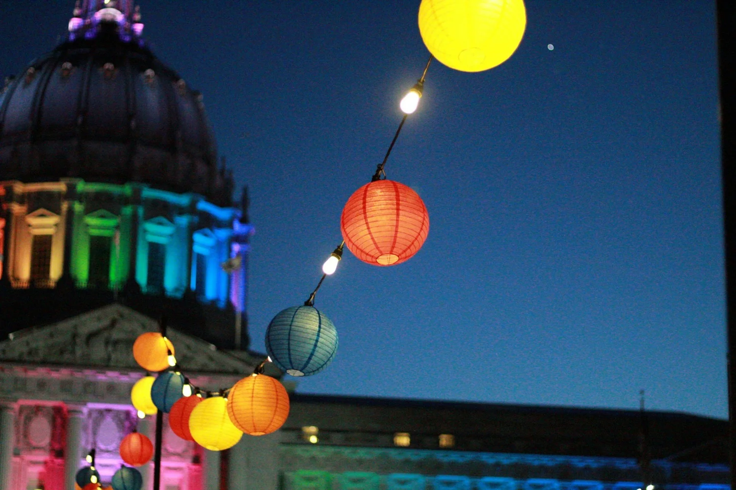 String of colorful paper lanterns at dusk in front of a domed building lit in rainbow colors