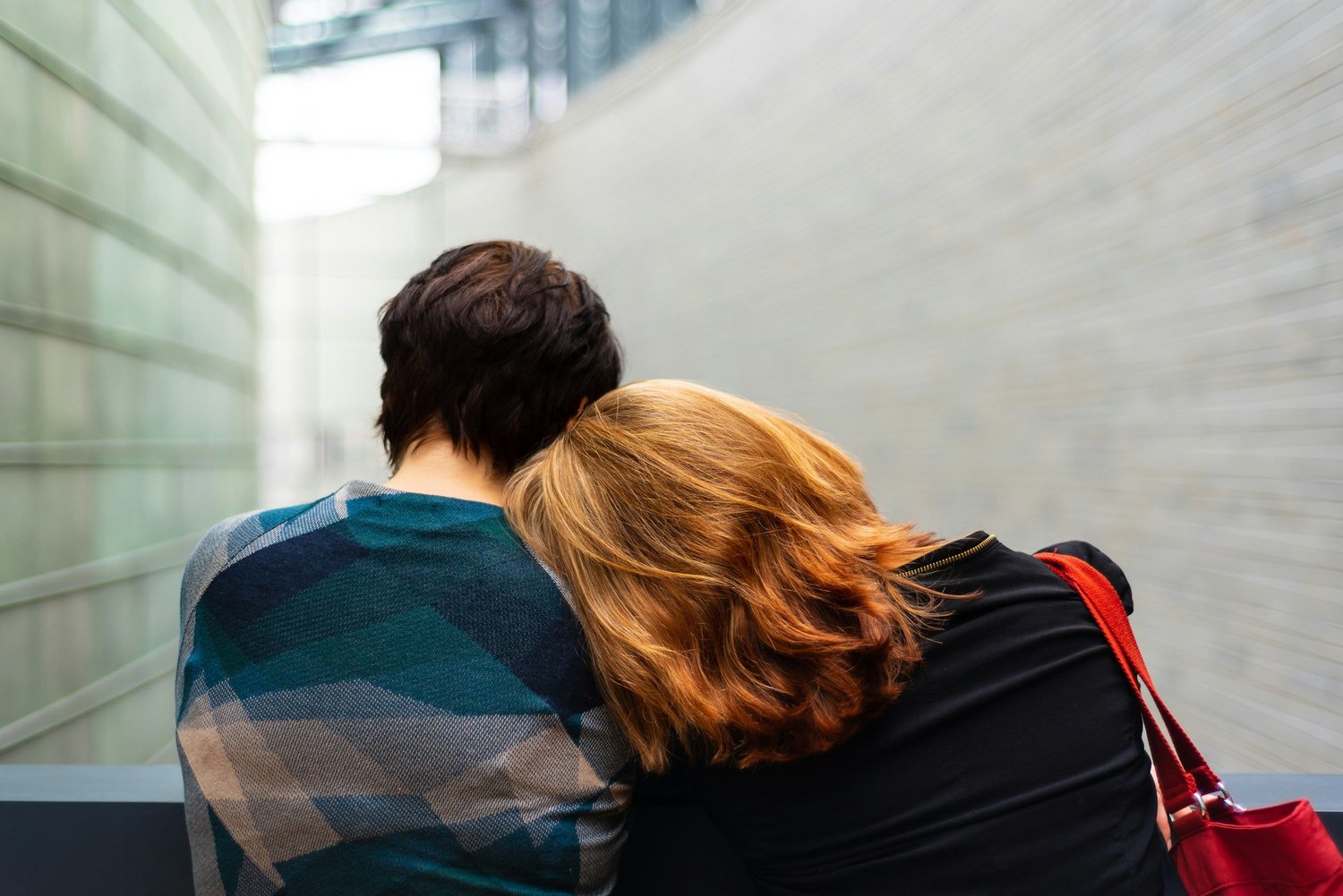 Two people seen from behind, one resting their head on the other's shoulder in a pale curved corridor