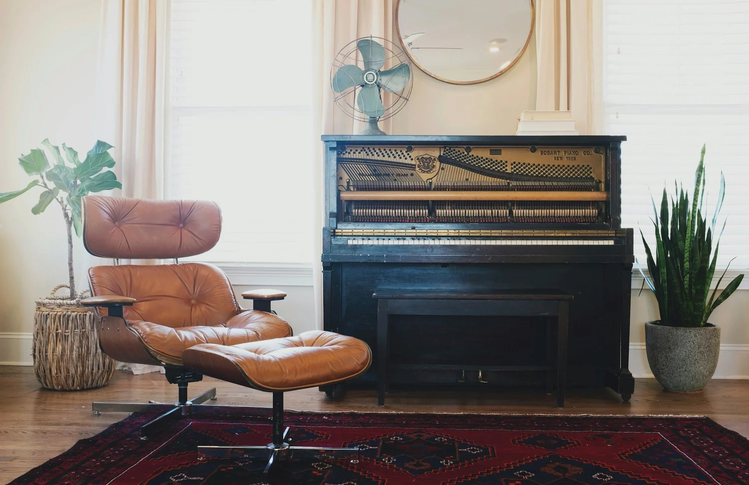 Leather lounge chair and ottoman beside an upright piano with a fan, round mirror and potted plants