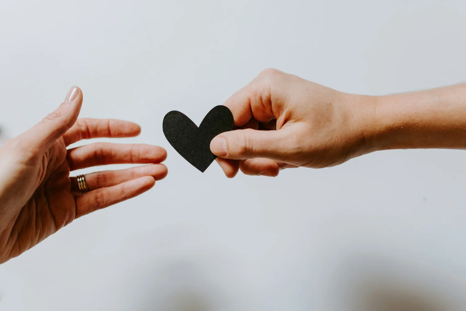 One hand passing a small black paper heart to another open hand against a pale background
