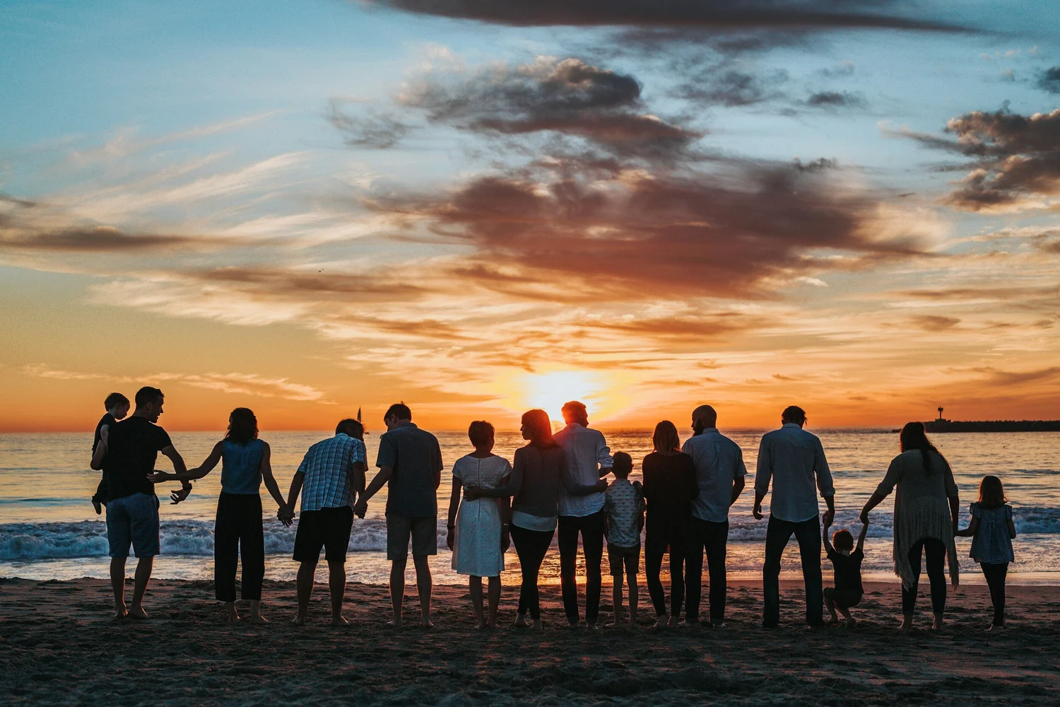 A row of people seen from behind holding hands on a beach, facing a sunset over the sea