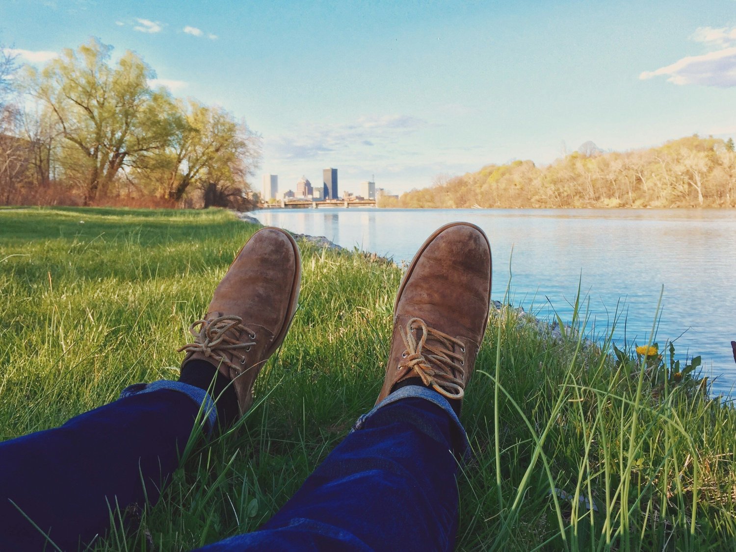 Legs in jeans and brown suede boots stretched out on grass beside a river with a city skyline beyond
