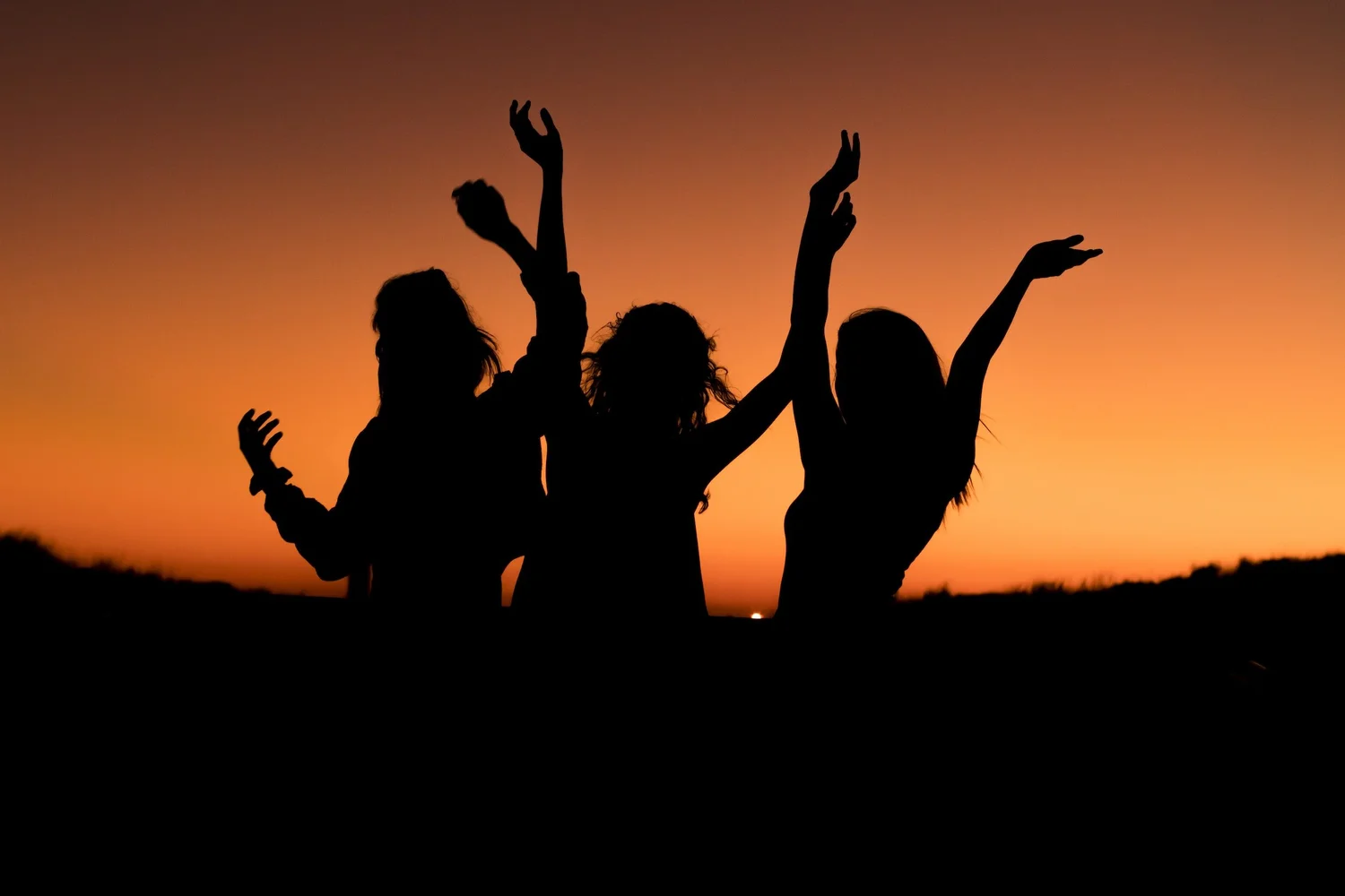 Silhouettes of three people with arms raised against an orange sunset sky