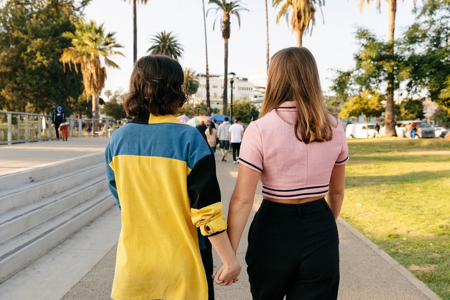 Two people seen from behind walking hand in hand along a sunny path lined with palm trees