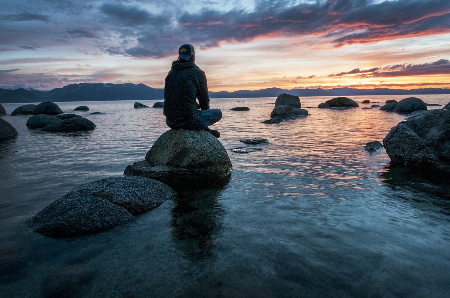 A person sits on a large rock in still water, facing mountains under a sunset sky