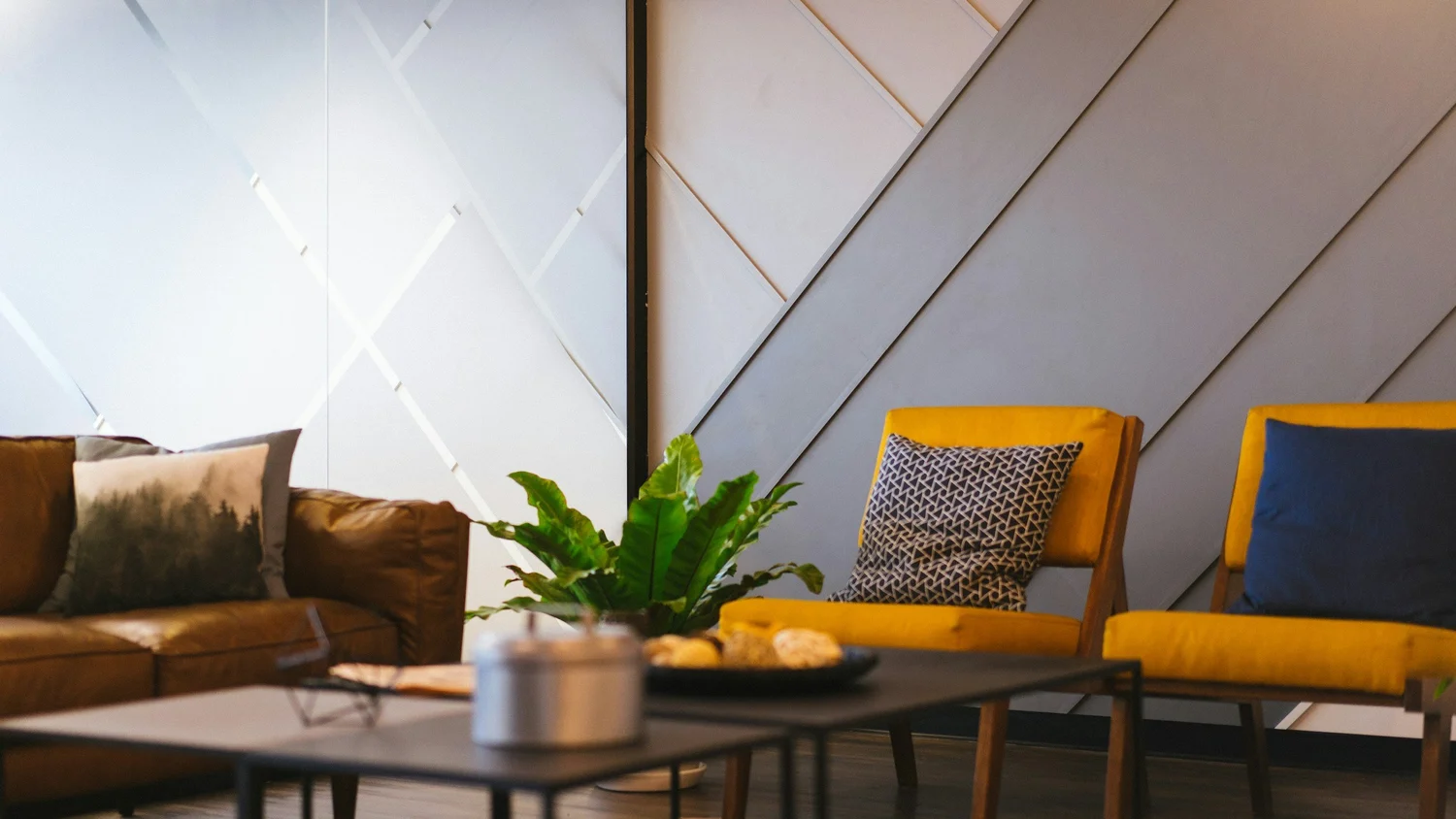 Lounge with a brown sofa, two yellow armchairs and a leafy plant on a low table by a gray paneled wall