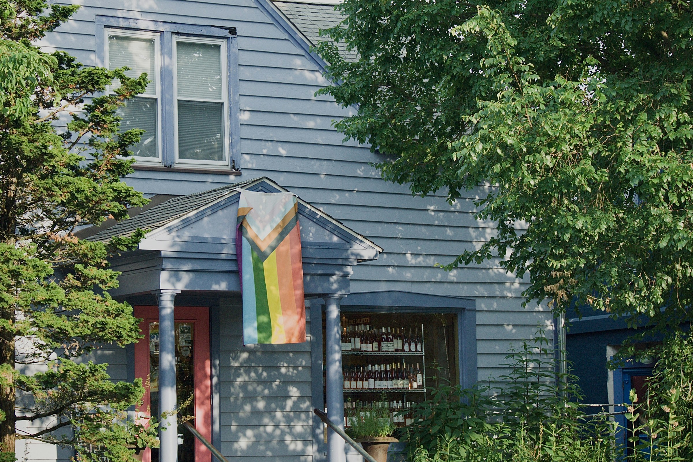 Progress Pride flag hanging from the front porch of a light blue clapboard house surrounded by green trees