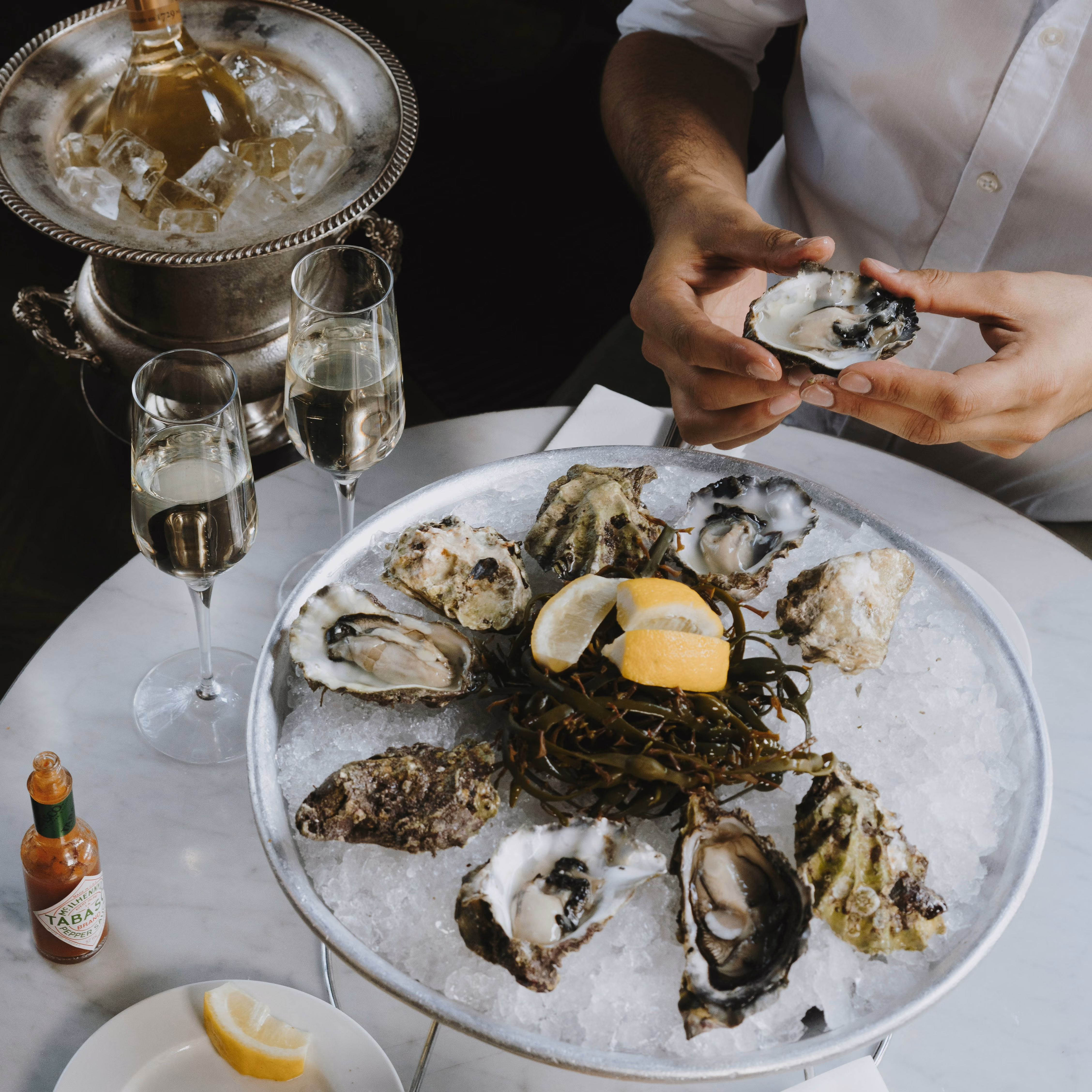 Person’s hands holding an opened oyster over a large metal tray filled with ice, oysters, seaweed, and lemon wedges on a round table with two glasses of white wine, a bottle of Tabasco sauce, and a wine bottle in an ice bucket.