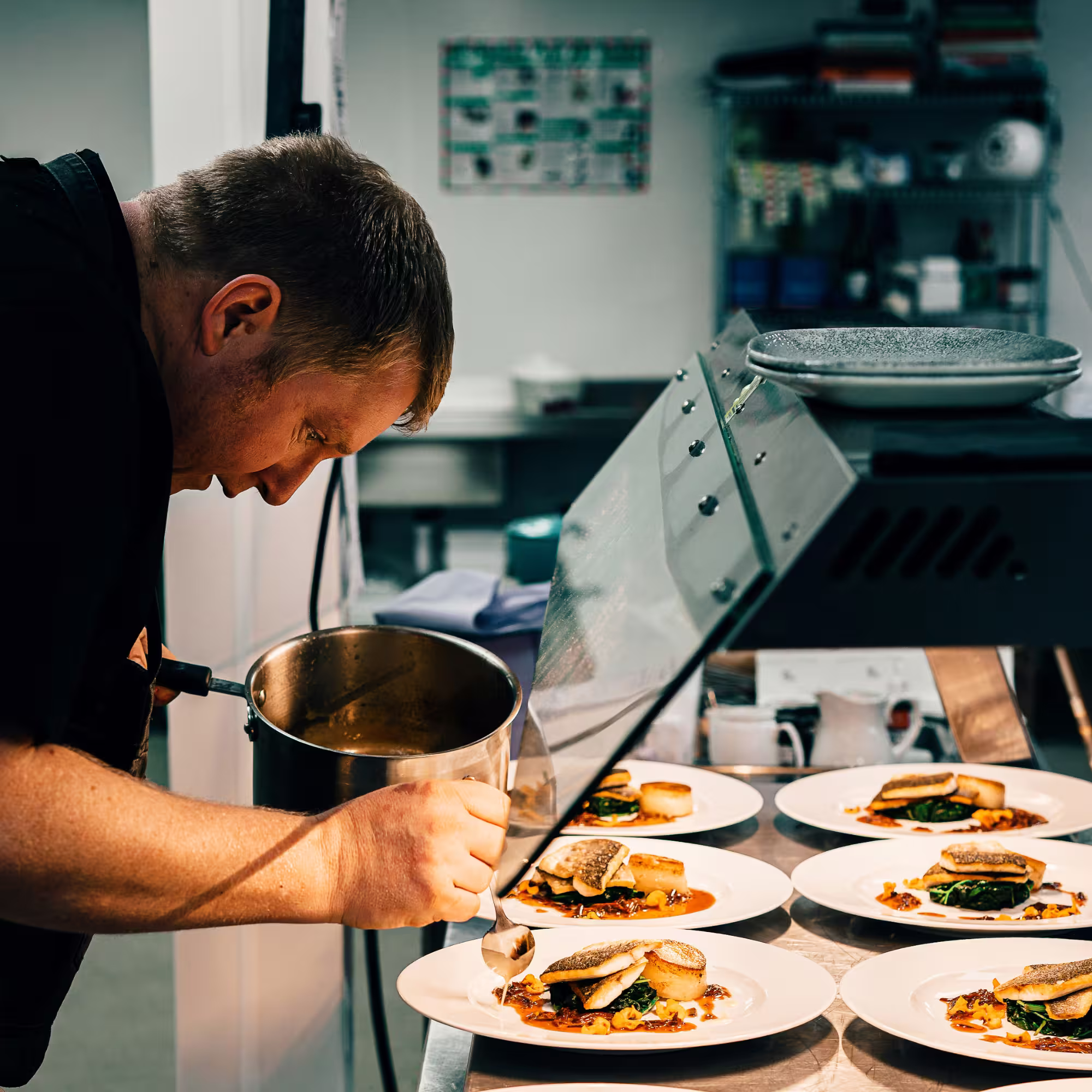 Chef carefully plating a gourmet dish with fish, greens, and sauce on white plates in a professional kitchen.