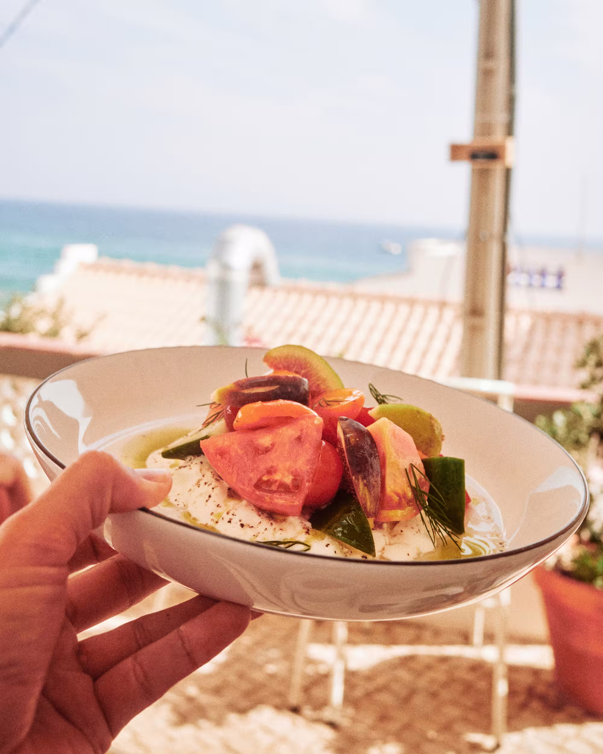 Hand holding a white bowl filled with fresh salad including tomato slices, cucumbers, olives, and herbs, with a sea view and rooftops in the blurred background.