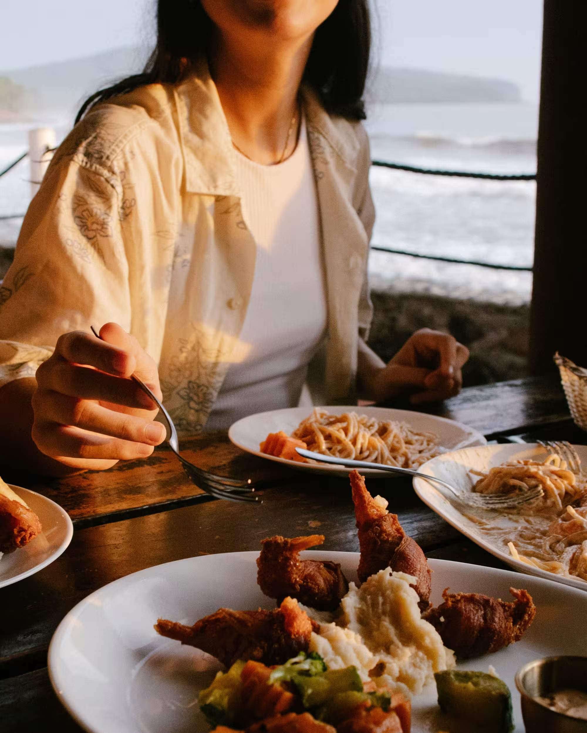Person in light-colored patterned shirt sitting at a wooden table by the sea, holding a fork near plates of food including fried shrimp and mashed potatoes, with waves and coastline in the background.