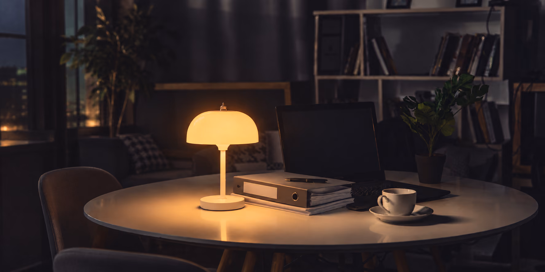 Dimly lit office with a round table featuring a glowing modern lamp, stacked binders, a laptop, and a white cup with saucer, surrounded by chairs and a background bookshelf and plant.