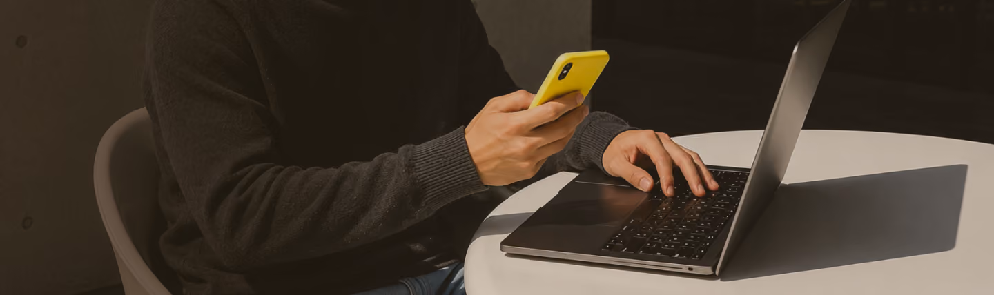 Person sitting at a round table using a laptop with one hand on the keyboard and holding a yellow smartphone in the other hand.