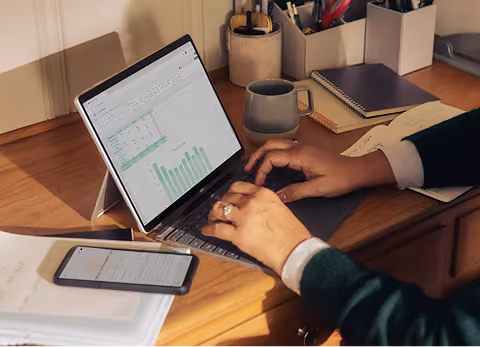 Person typing on a laptop showing a spreadsheet with charts, a phone and notebook on the desk
