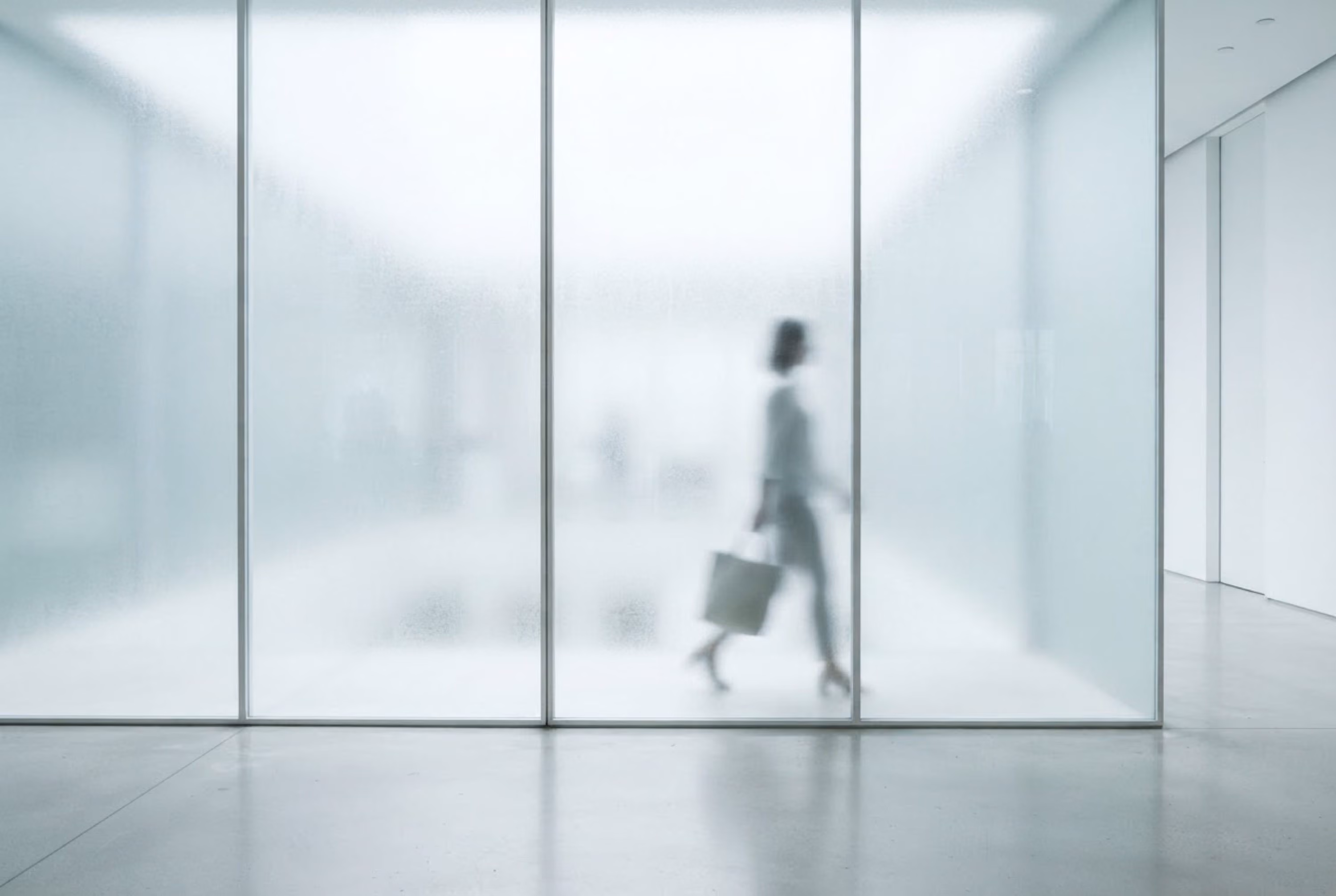 Verschwommene Silhouette einer Person, die in einem modernen, hellen Raum mit großen Glaswänden und reflektierendem Boden geht und eine Tasche trägt.