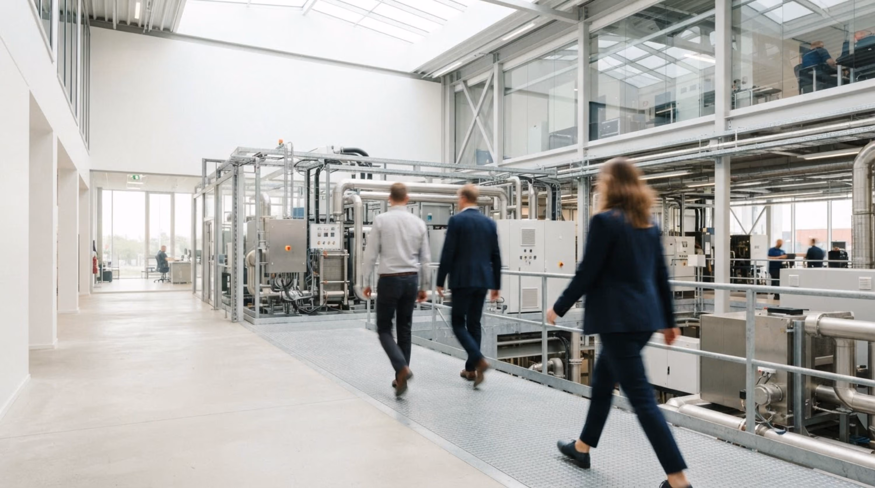 Drei Personen im Geschäftskleid gehen in einer modernen Industrieanlage mit großen Maschinen und Rohren entlang eines Metallgitterwegs.