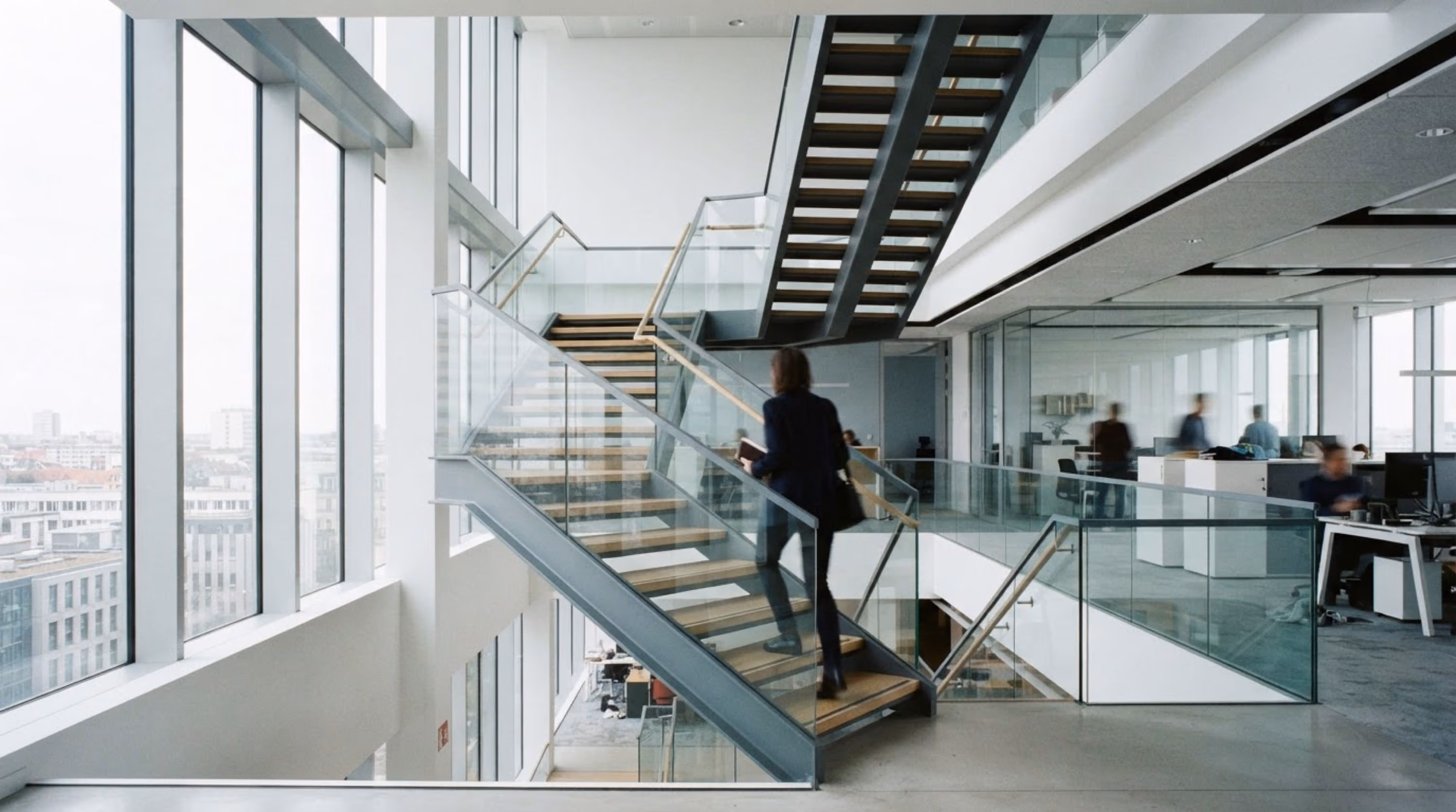 Frau steigt in modernem Bürogebäude eine Treppe mit Glasgeländer hinauf, umgeben von großen Fenstern und offenen Arbeitsbereichen.