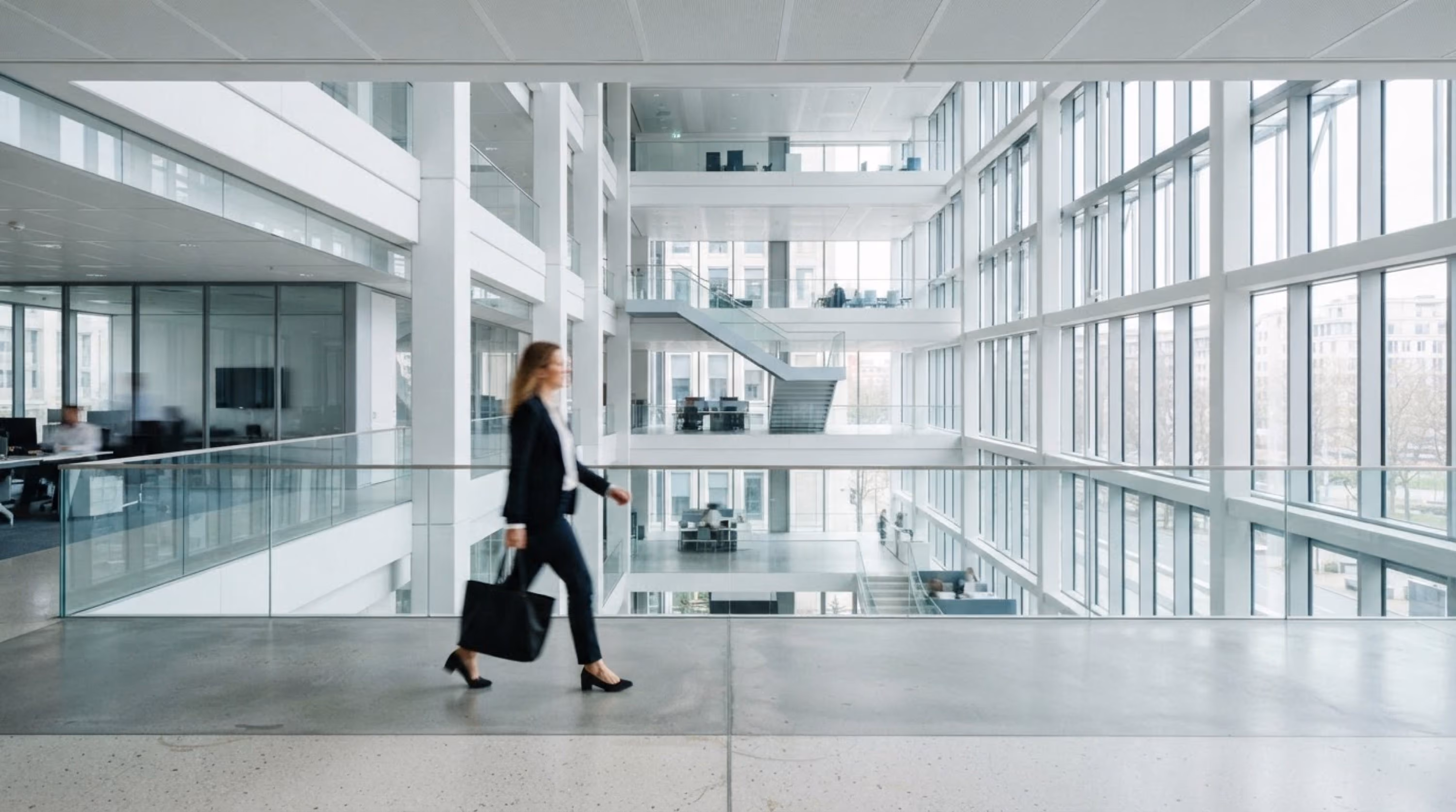 Eine Frau in Geschäftskleidung geht mit einer Tasche durch ein modernes, helles Bürogebäude mit großen Fenstern und mehrstöckigen Arbeitsbereichen.