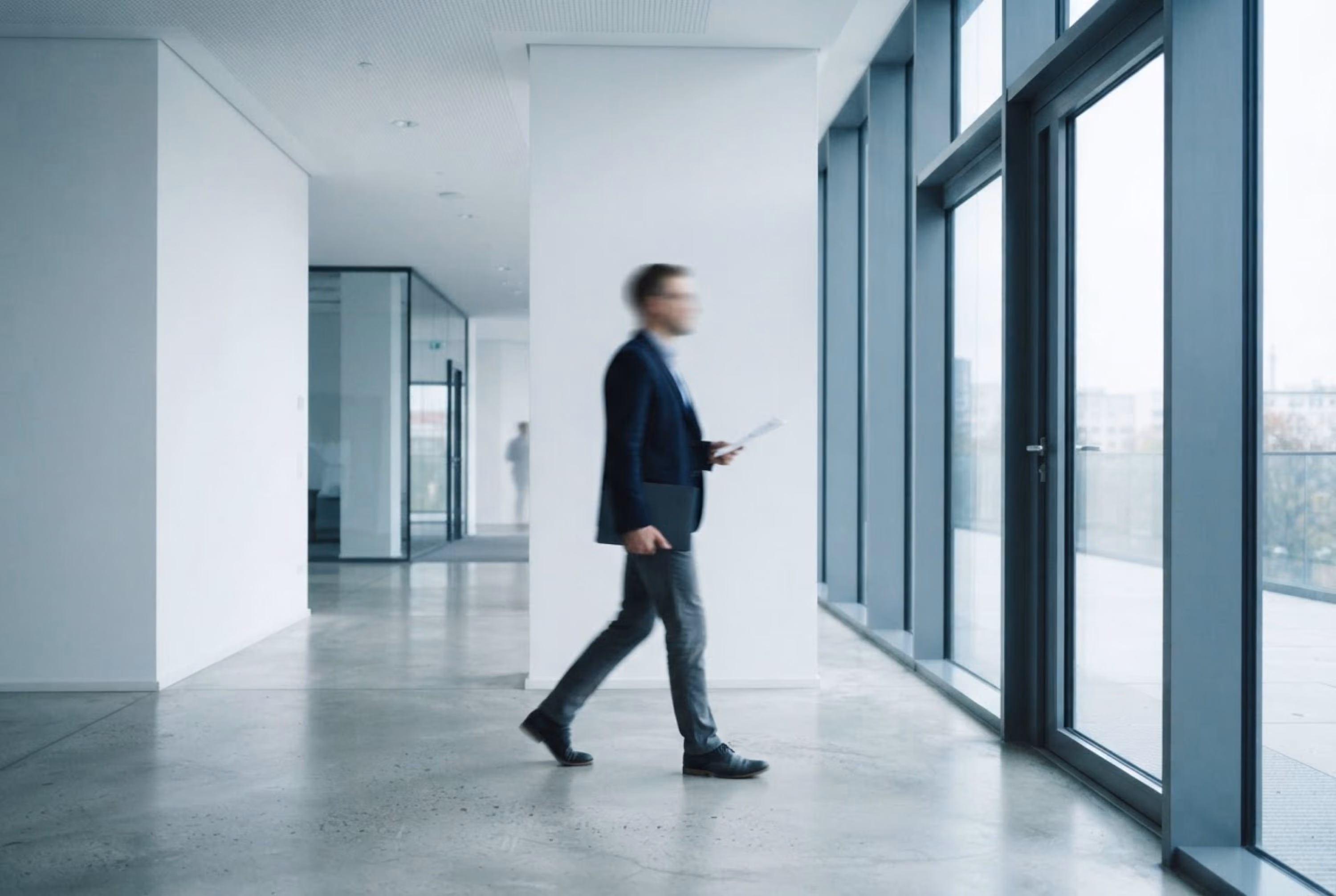 Ein unscharf abgebildeter Mann in Businesskleidung, der in einem modernen Bürogebäude am Fenster entlanggeht und in der Hand ein Tablet hält.