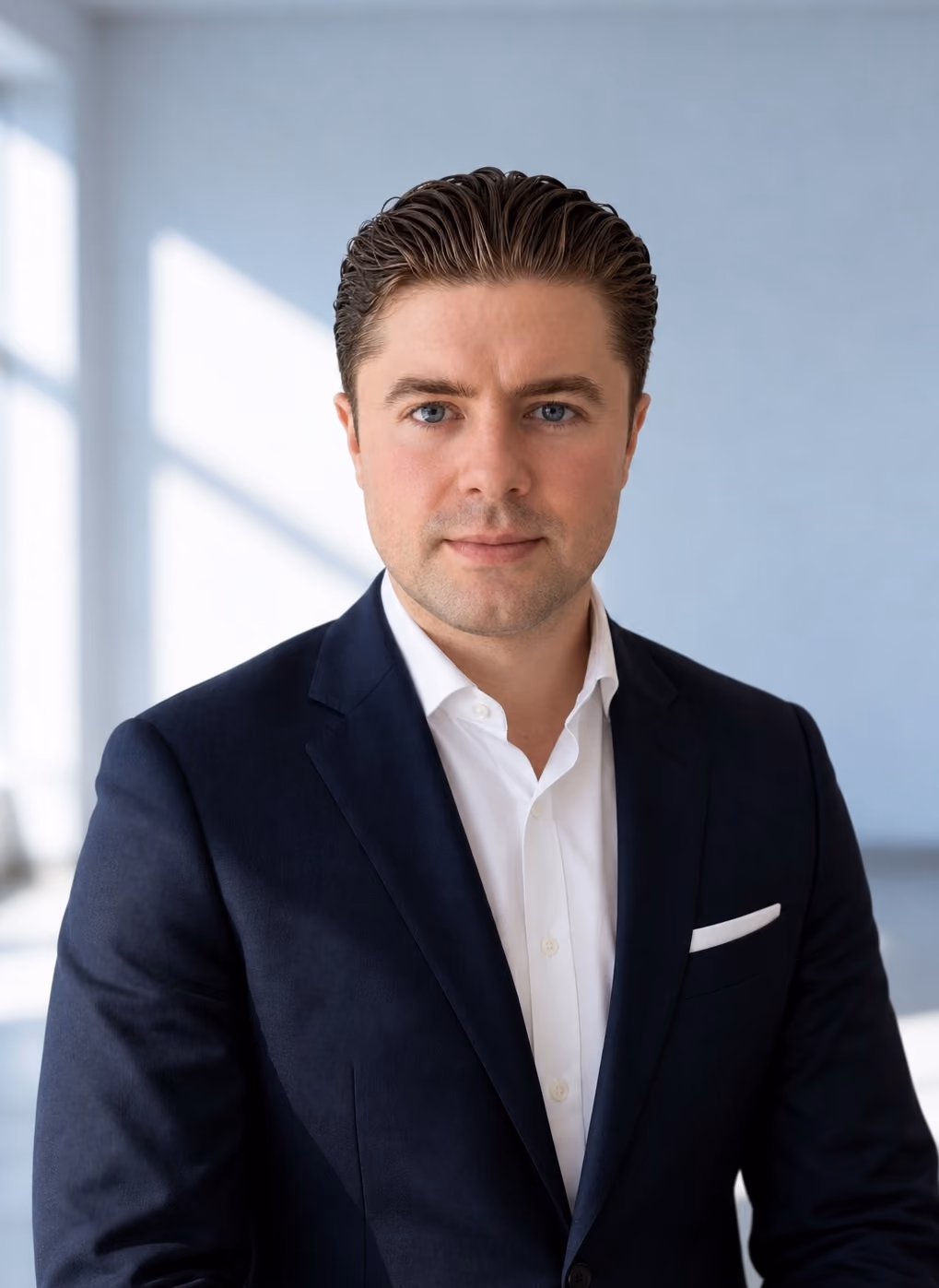 Porträt eines Mannes mit hellen blauen Augen, dunklem, zurückgekämmtem Haar, der einen dunklen Anzug mit weißem Hemd und Einstecktuch trägt, vor einem hellen unscharfen Hintergrund mit großen Fenstern.