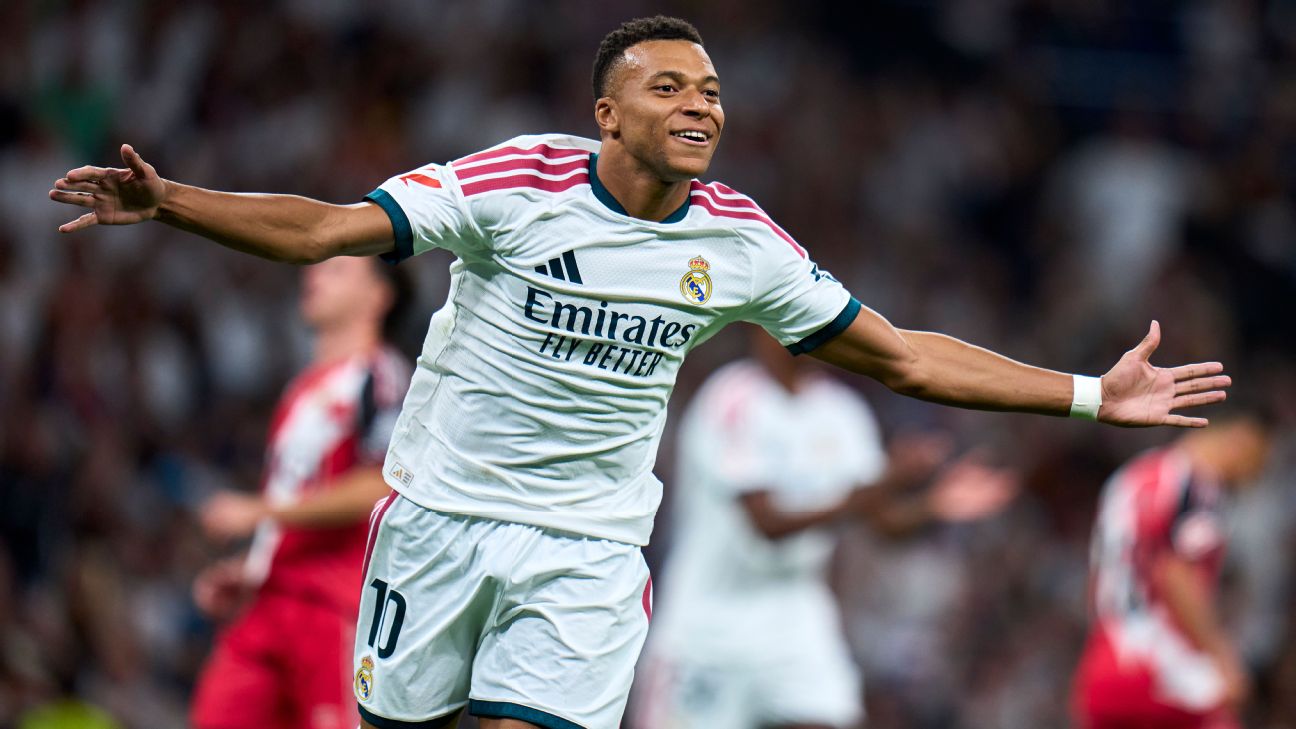 Kylian Mbappé celebrates with both arms outstretched and a broad smile in Real Madrid's white home shirt, with Rayo Vallecano players in red and white blurred behind him at the Santiago Bernabéu.