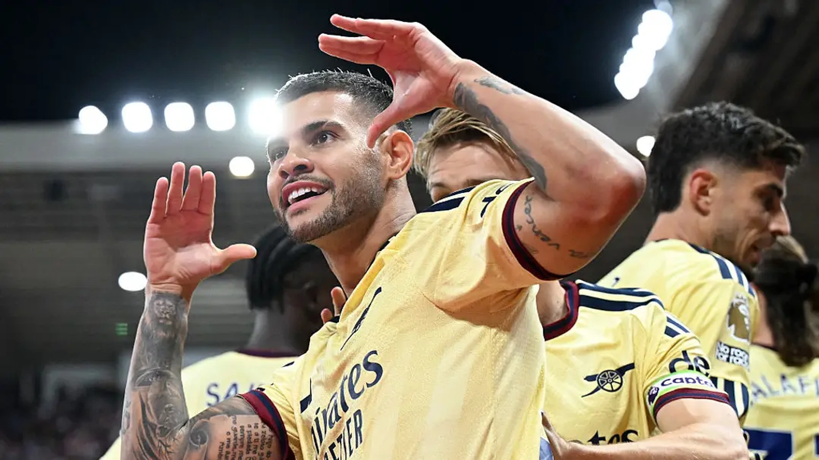 Bruno Guimarães in Arsenal's yellow away shirt celebrates with both hands raised, teammates behind him under the floodlights.