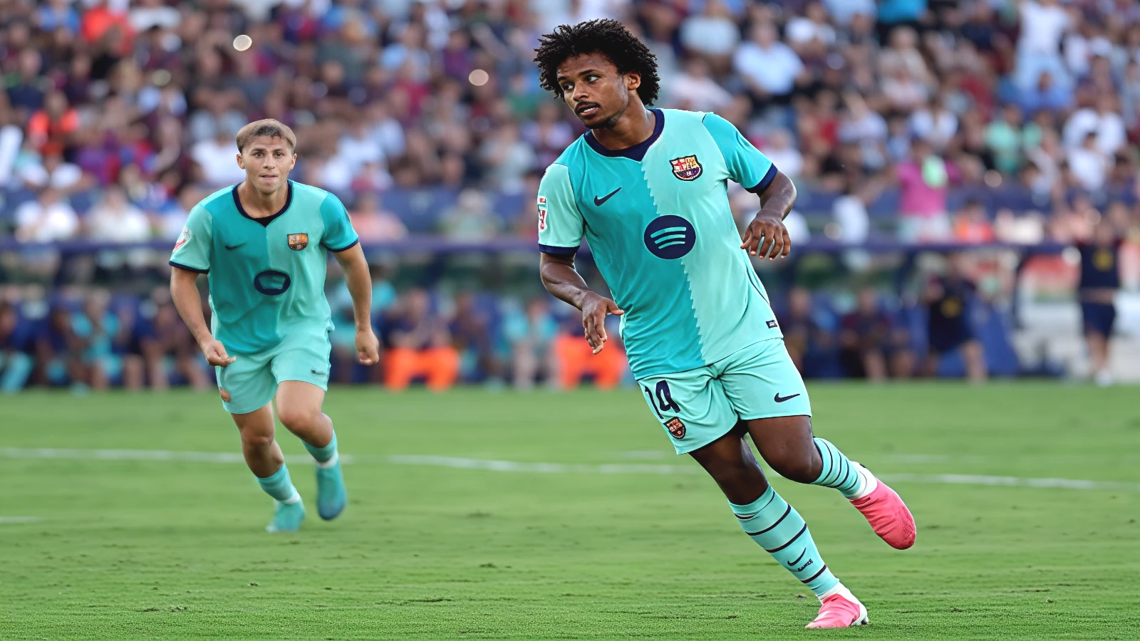 Karim Adeyemi runs forward in Barcelona's turquoise away kit at the Ciutat de València, a teammate following behind him and Levante supporters filling the stand.