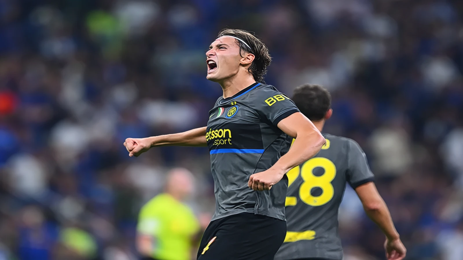 Inter player in the grey kit shouts in celebration with both fists clenched, a teammate wearing number 28 behind him and a packed stand out of focus in the background