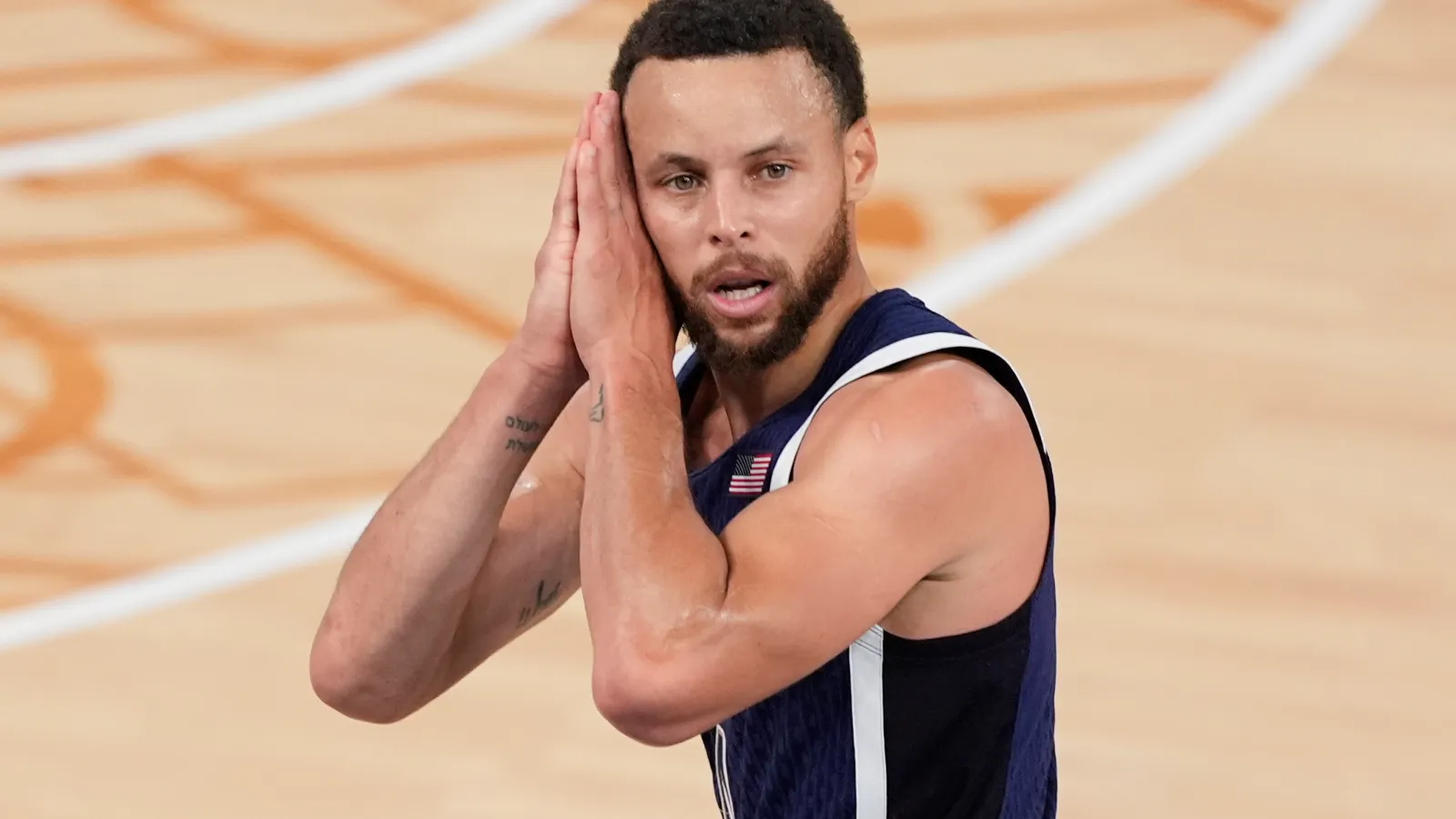 Stephen Curry presses his palms together beside his head, wearing a navy USA jersey on a basketball court.