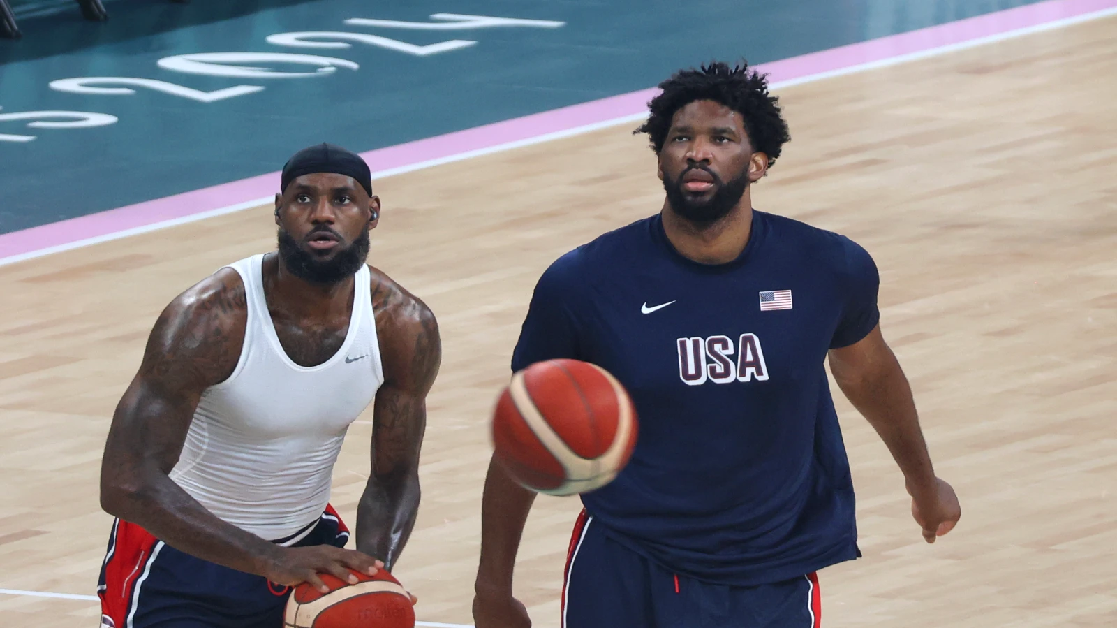 LeBron James in a white sleeveless top holds a basketball on a practice court beside Joel Embiid in a navy USA shirt, with 2024 markings on the floor behind them.