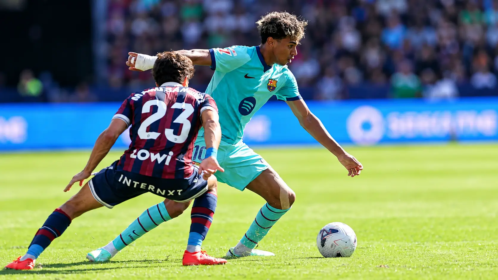 Lamine Yamal, in Barcelona's turquoise away kit with the number 10 on his shorts, shapes to go past an opponent in a blue and red striped number 23 shirt, the ball at his feet and a packed stand behind them.