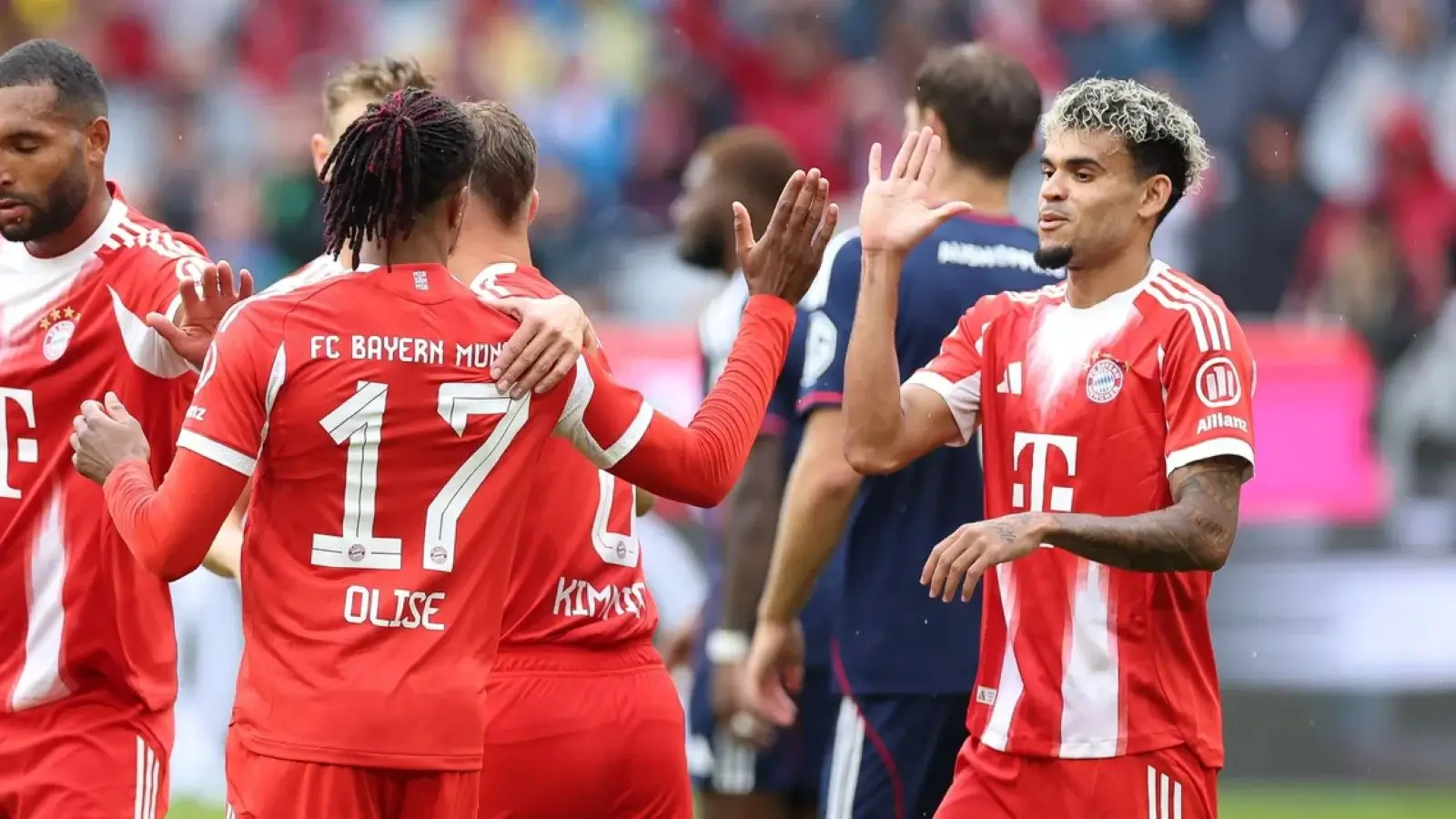 Michael Olise is embraced by a team-mate as Luis Díaz raises a hand to celebrate with him, all three in Bayern Munich's red home shirts