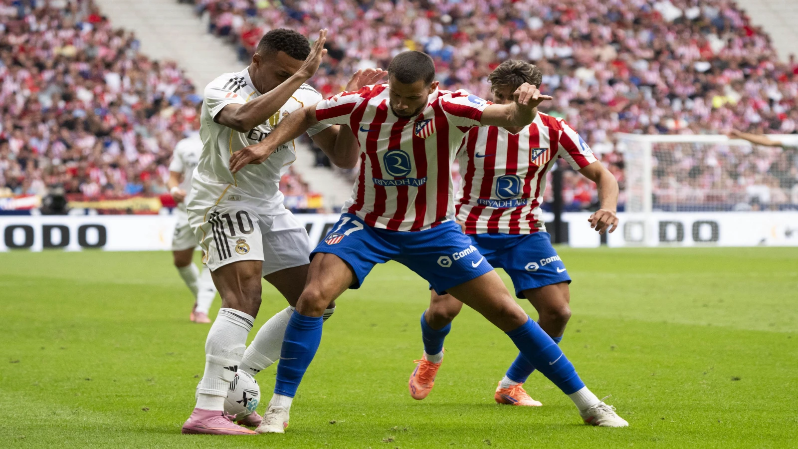 A Real Madrid player in white, wearing number 10, shields the ball from an Atlético Madrid player in red and white stripes, while a second Atlético player closes in behind, in front of a packed crowd.