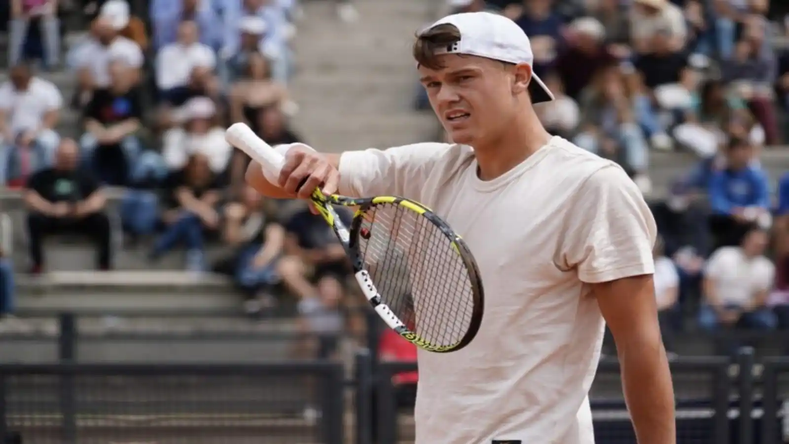 Holger Rune holds his racket up by the frame on a clay court, in a plain t-shirt and a backwards white cap