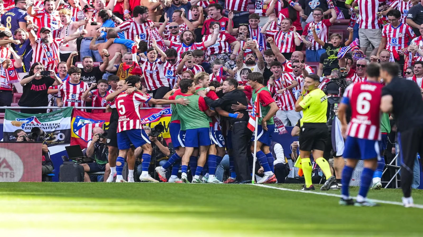 Atlético Madrid players and substitutes crowd around their manager by the corner flag to celebrate a goal, with the home supporters standing and cheering behind them.