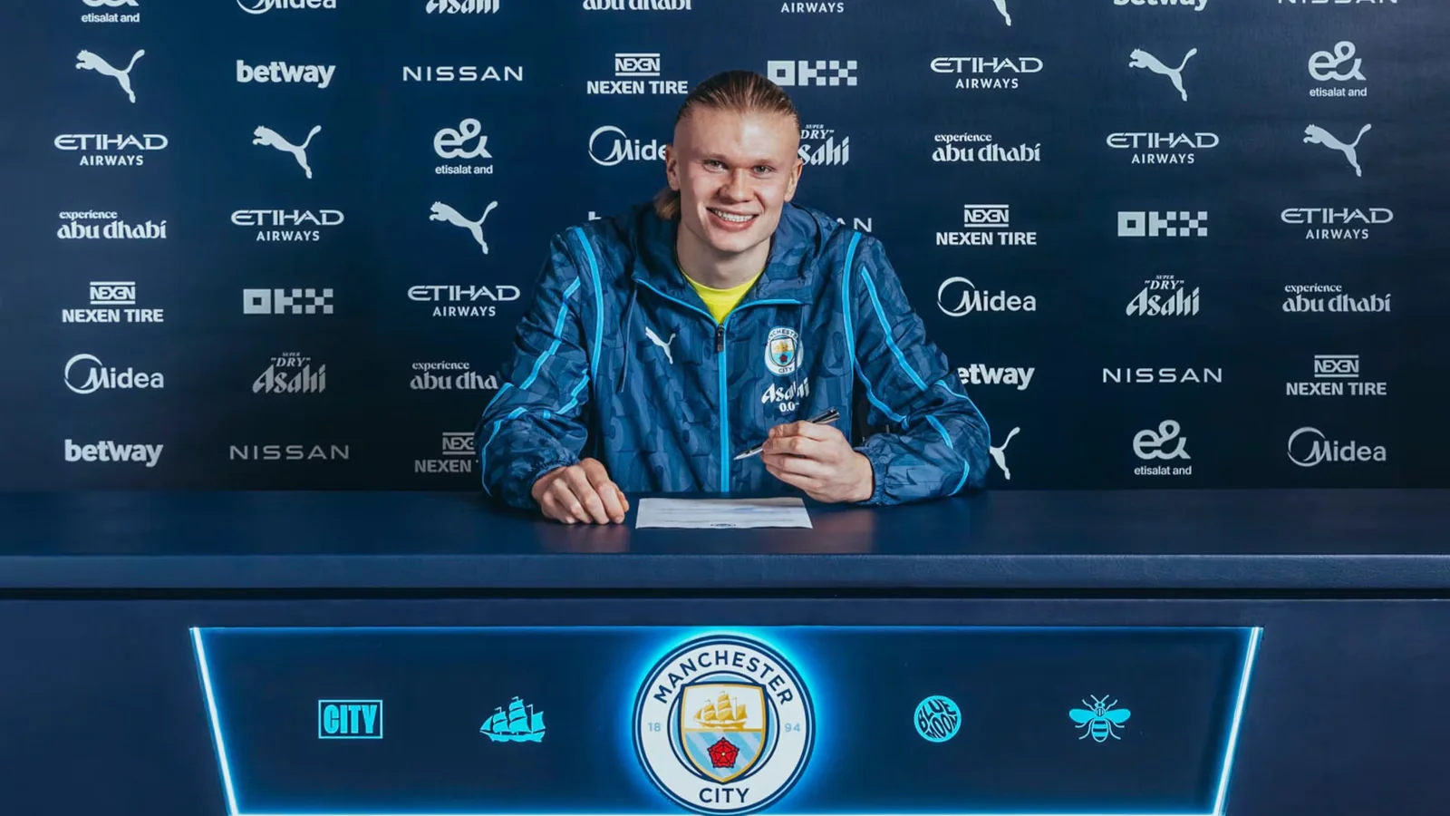 Erling Haaland smiles while signing his Manchester City contract at a table bearing the club crest.