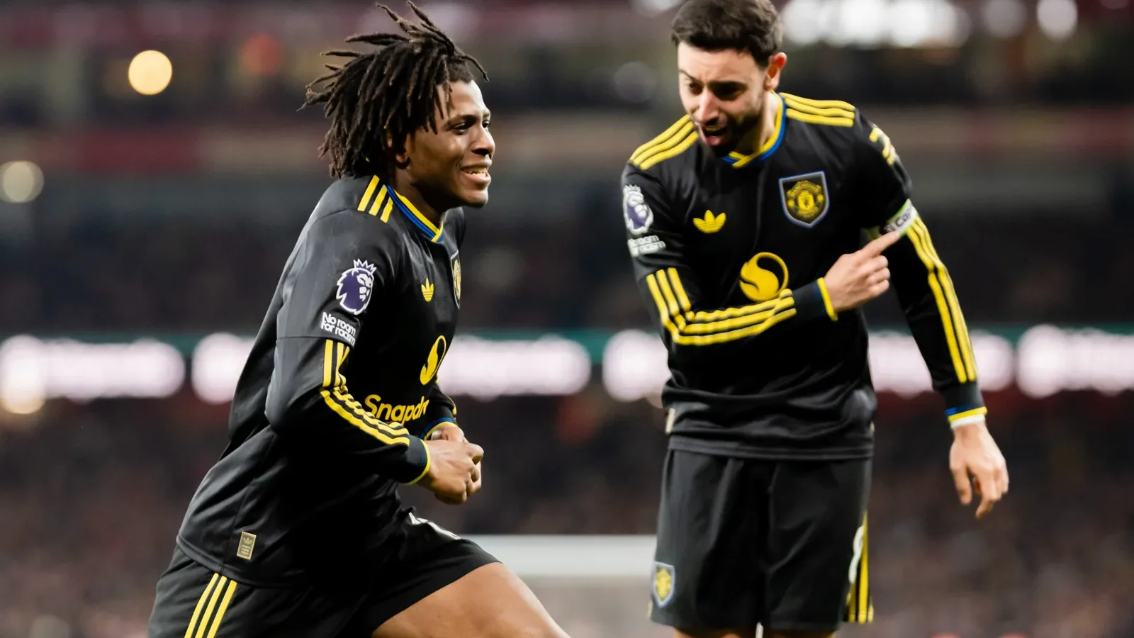 Patrick Dorgu smiles alongside Bruno Fernandes as the Manchester United teammates wear black kits during a match.