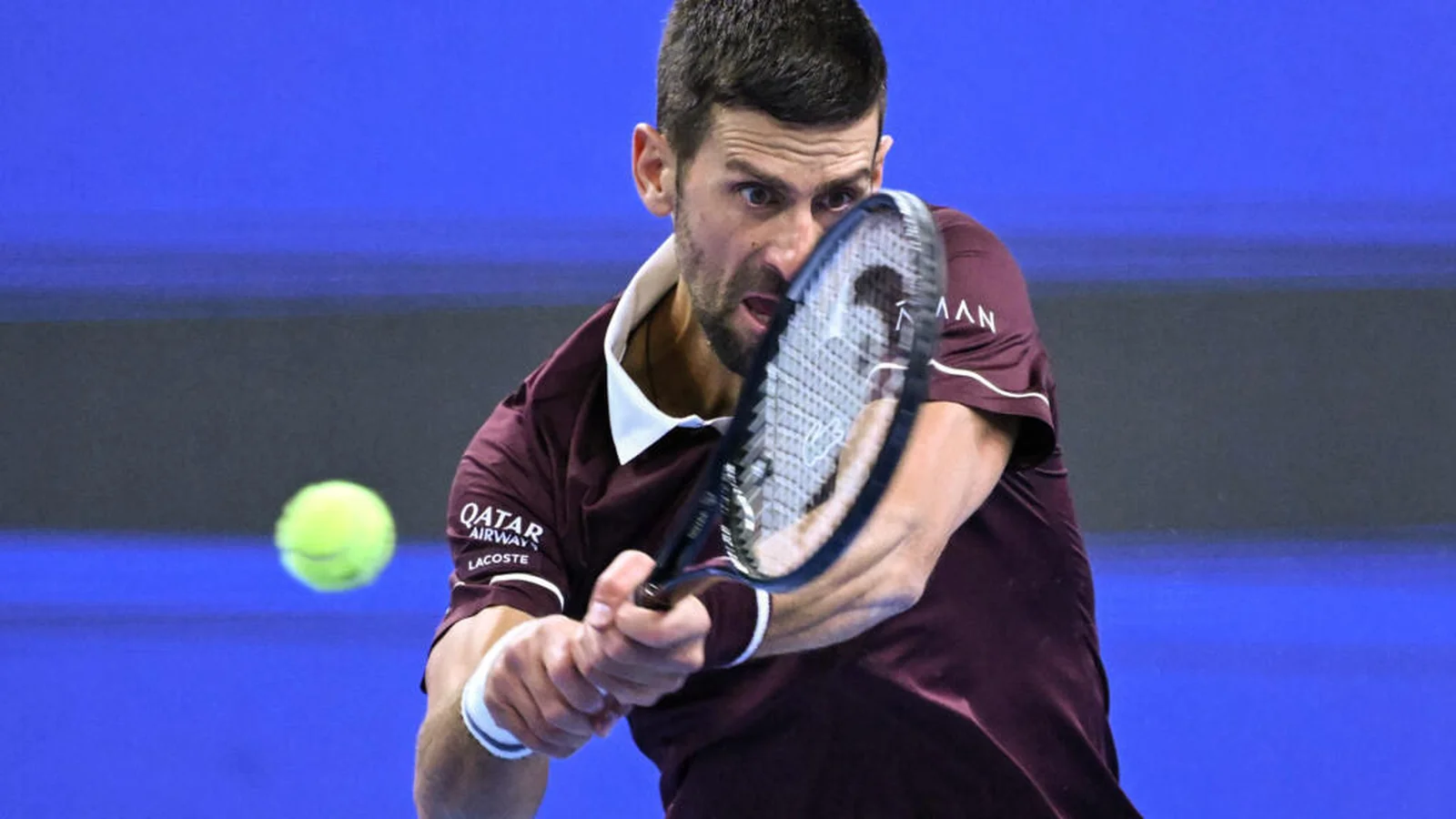 Novak Djokovic hitting a two-handed backhand while wearing a burgundy shirt.
