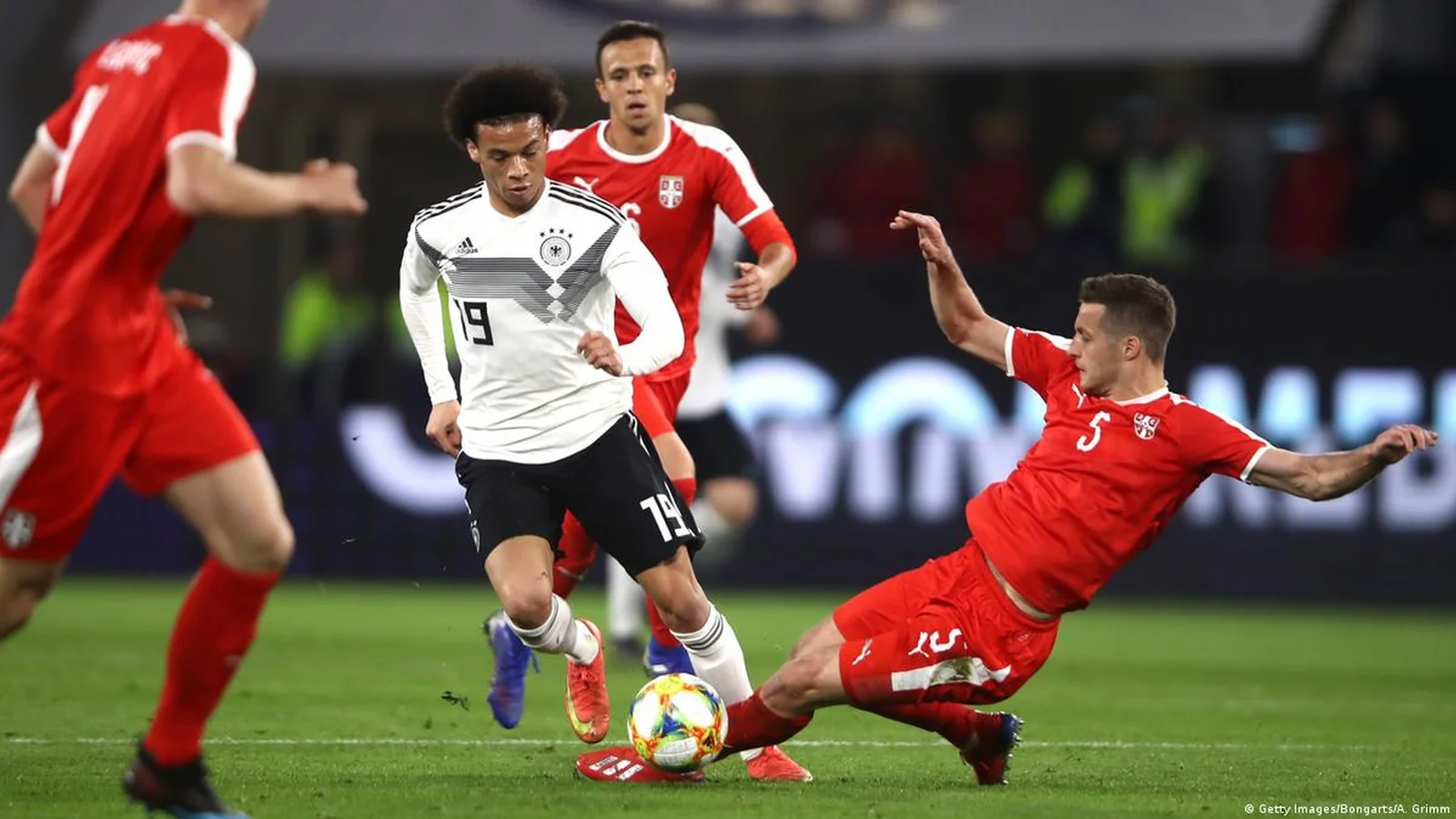 Germany player in a white jersey dribbles as a Serbia defender slides in to tackle.