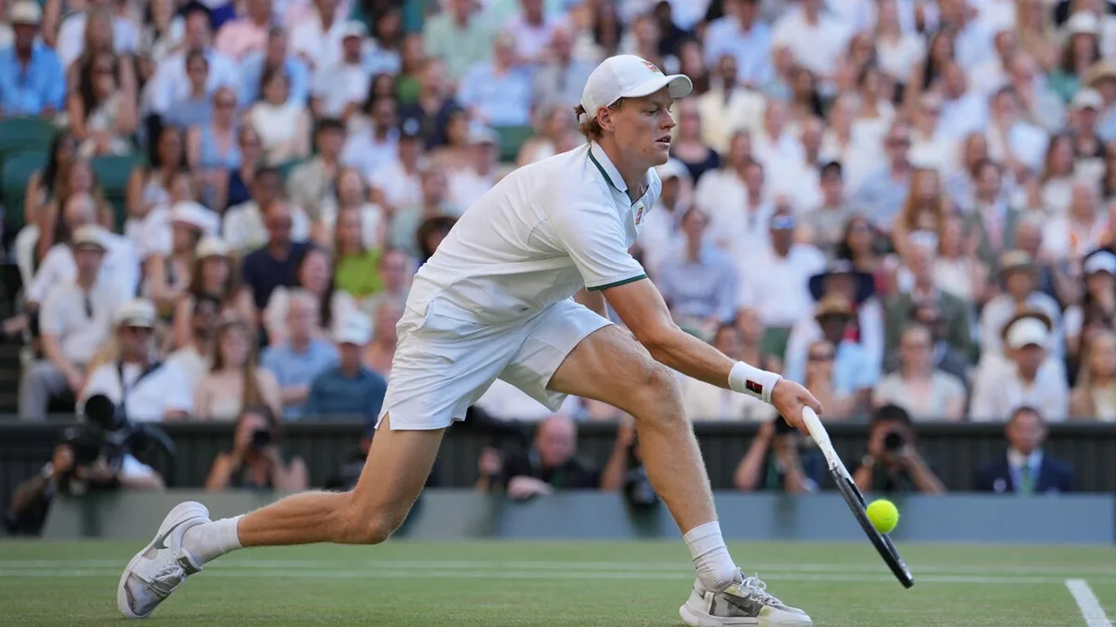 Jannik Sinner stretches to return a low ball on a grass tennis court, wearing white kit and a cap.
