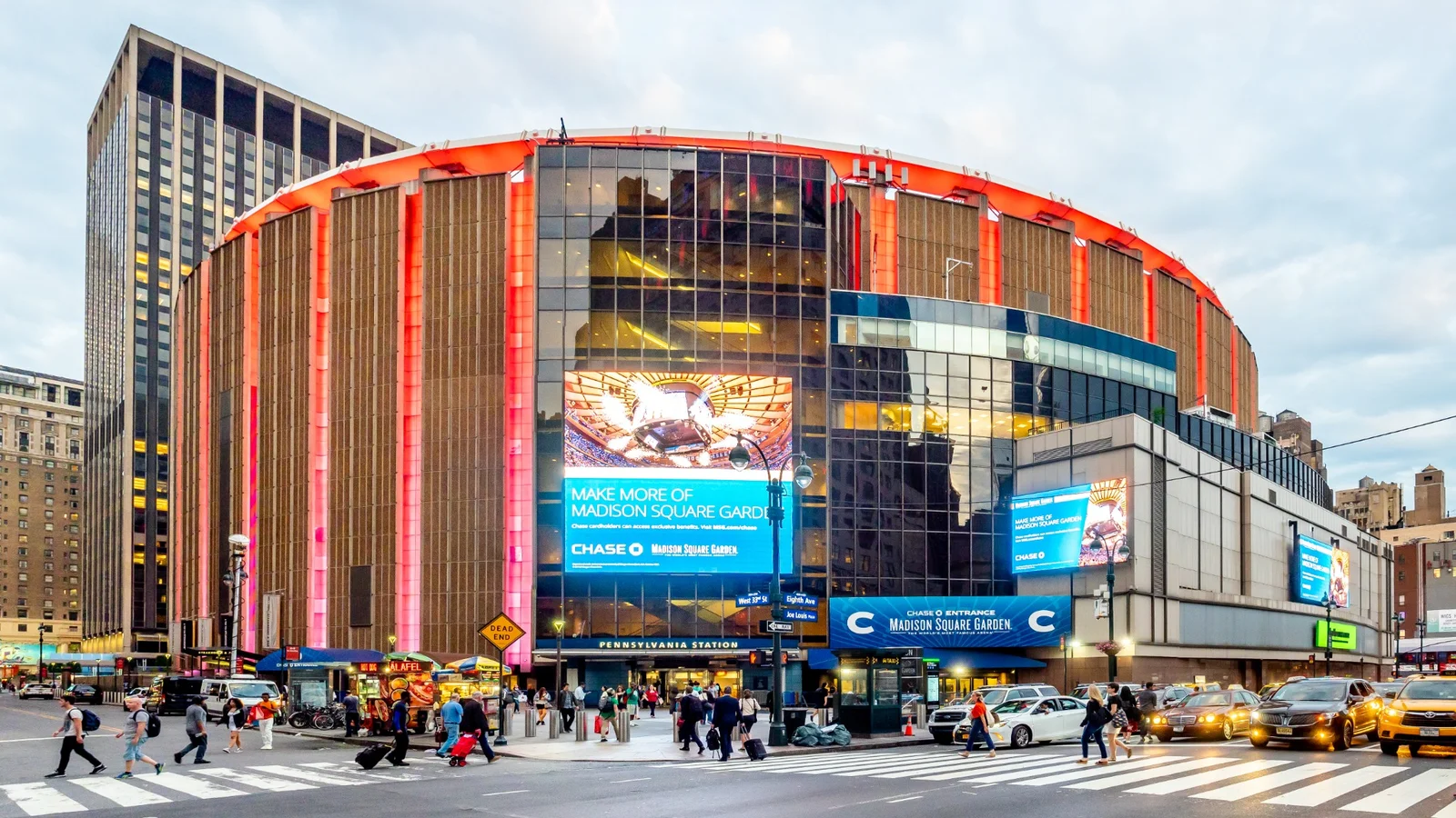 Exterior of Madison Square Garden in New York City, with illuminated red facade, pedestrians and traffic outside.