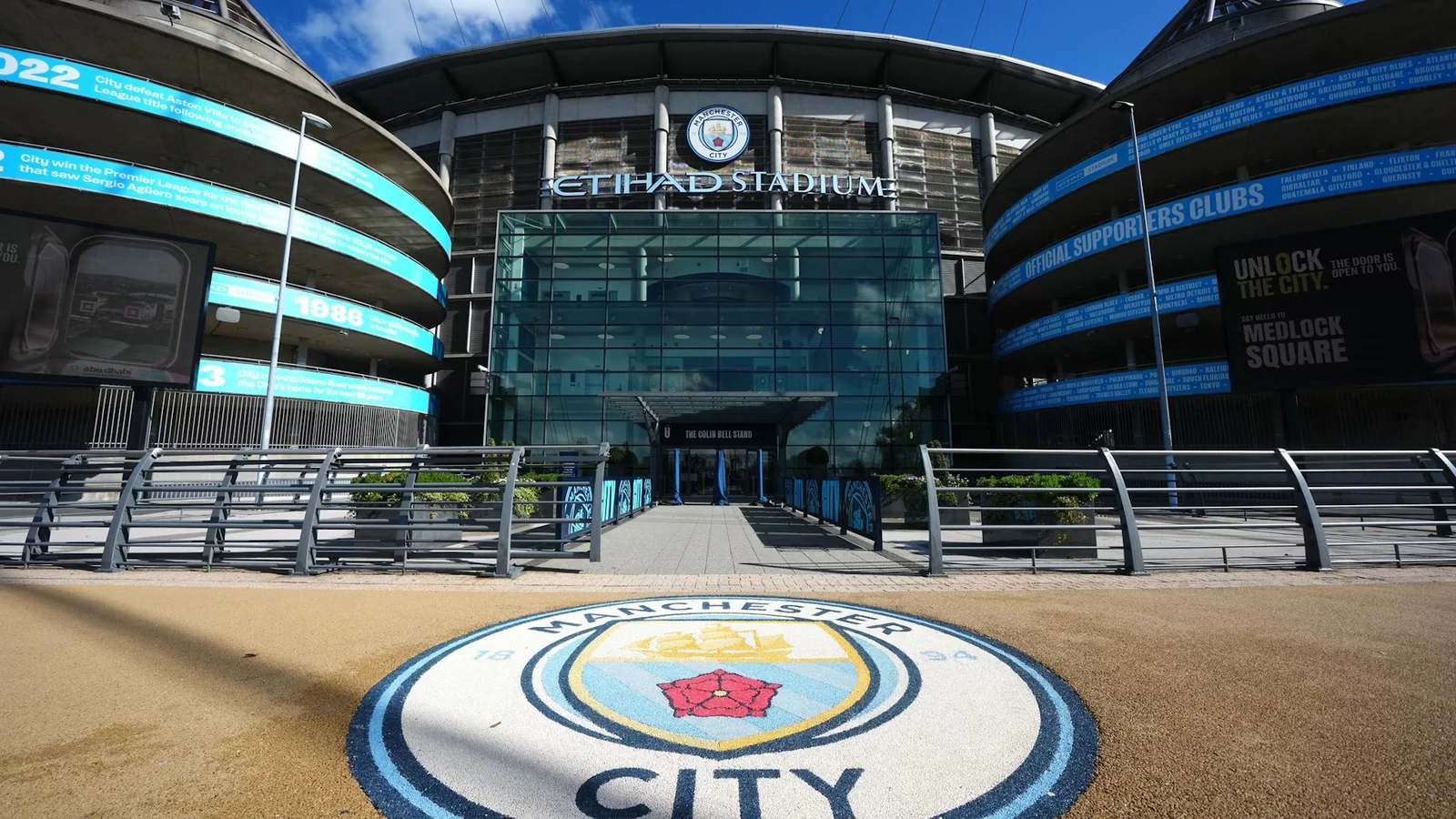 Exterior view of Manchester City’s Etihad Stadium with the club crest displayed in the foreground.