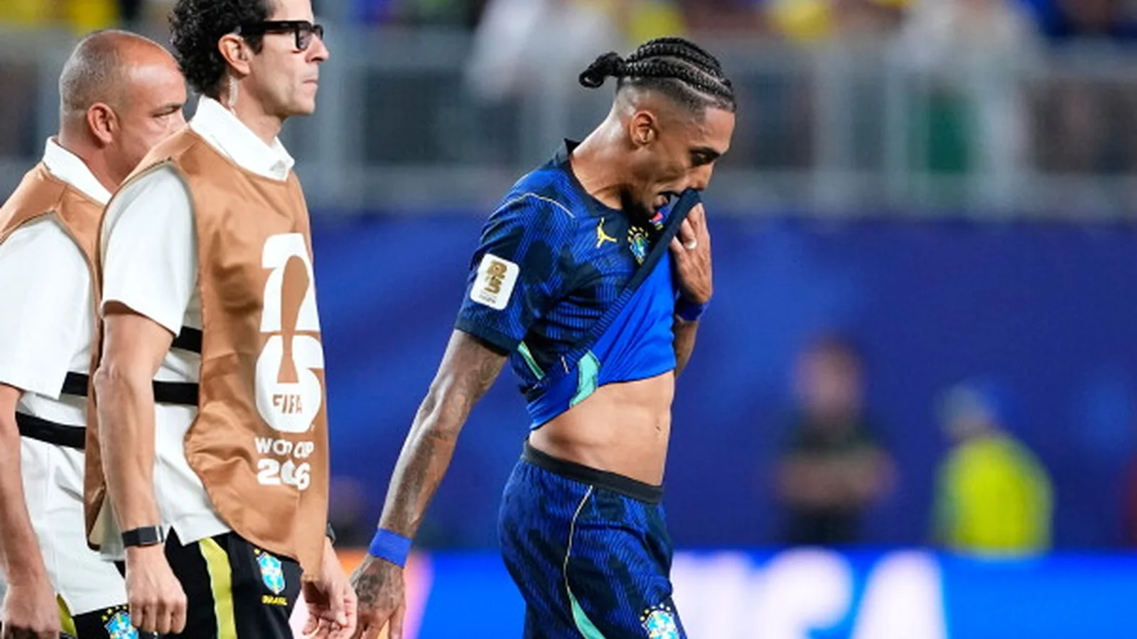 Raphinha walks beside Brazil team staff, holding his blue national-team shirt to his mouth.