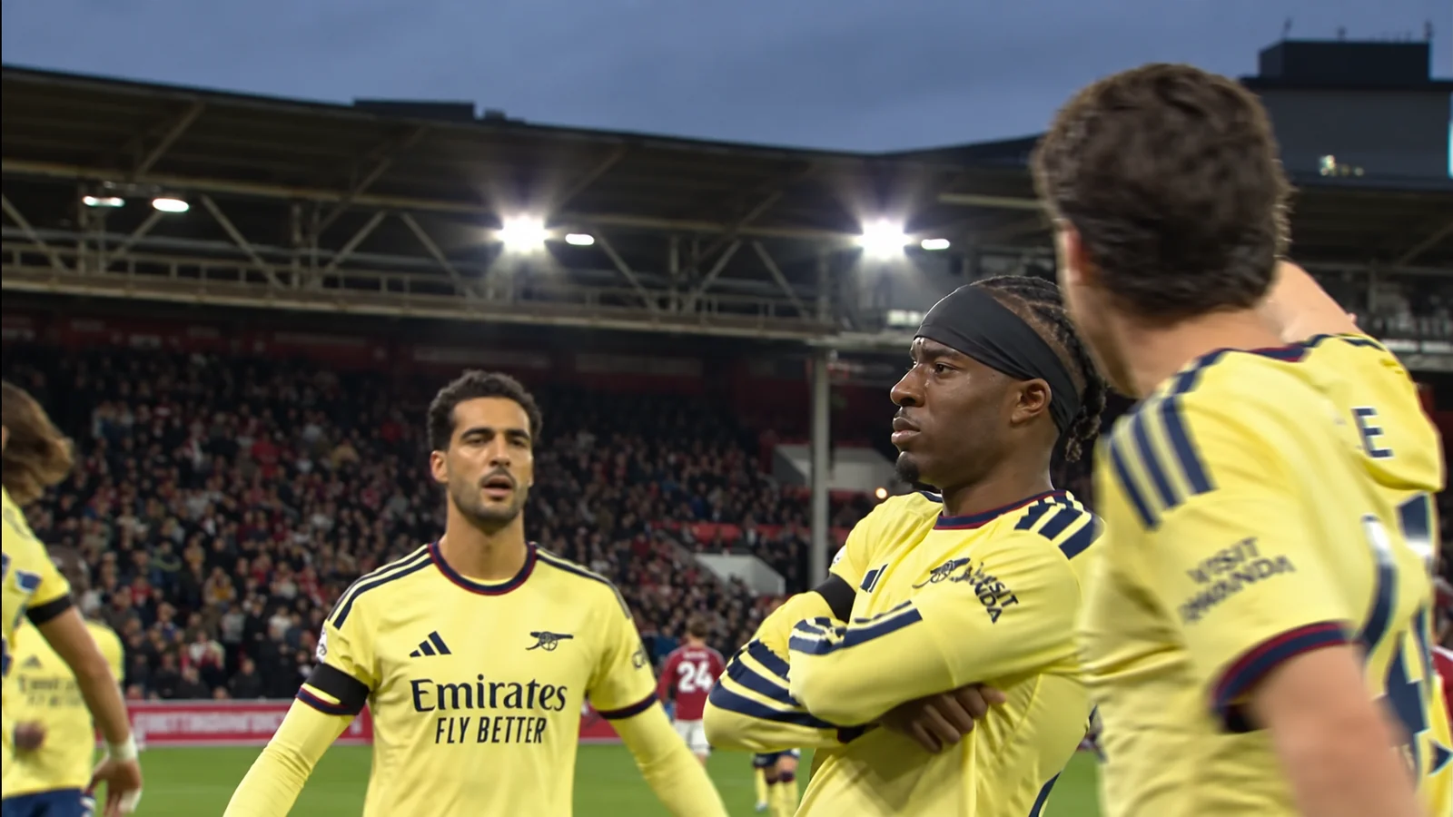 Arsenal players in yellow away kits gather on the pitch under stadium floodlights.