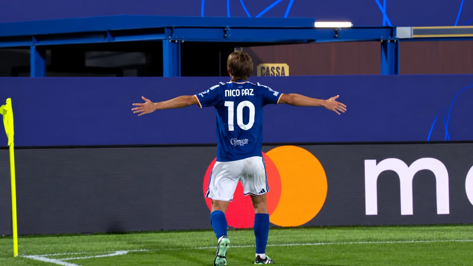 Nico Paz celebrates near the corner flag with his arms outstretched during a Champions League match.